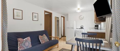 A compact living space with a sofa, dining table, chairs, a TV, kitchen area, and a patterned rug.
