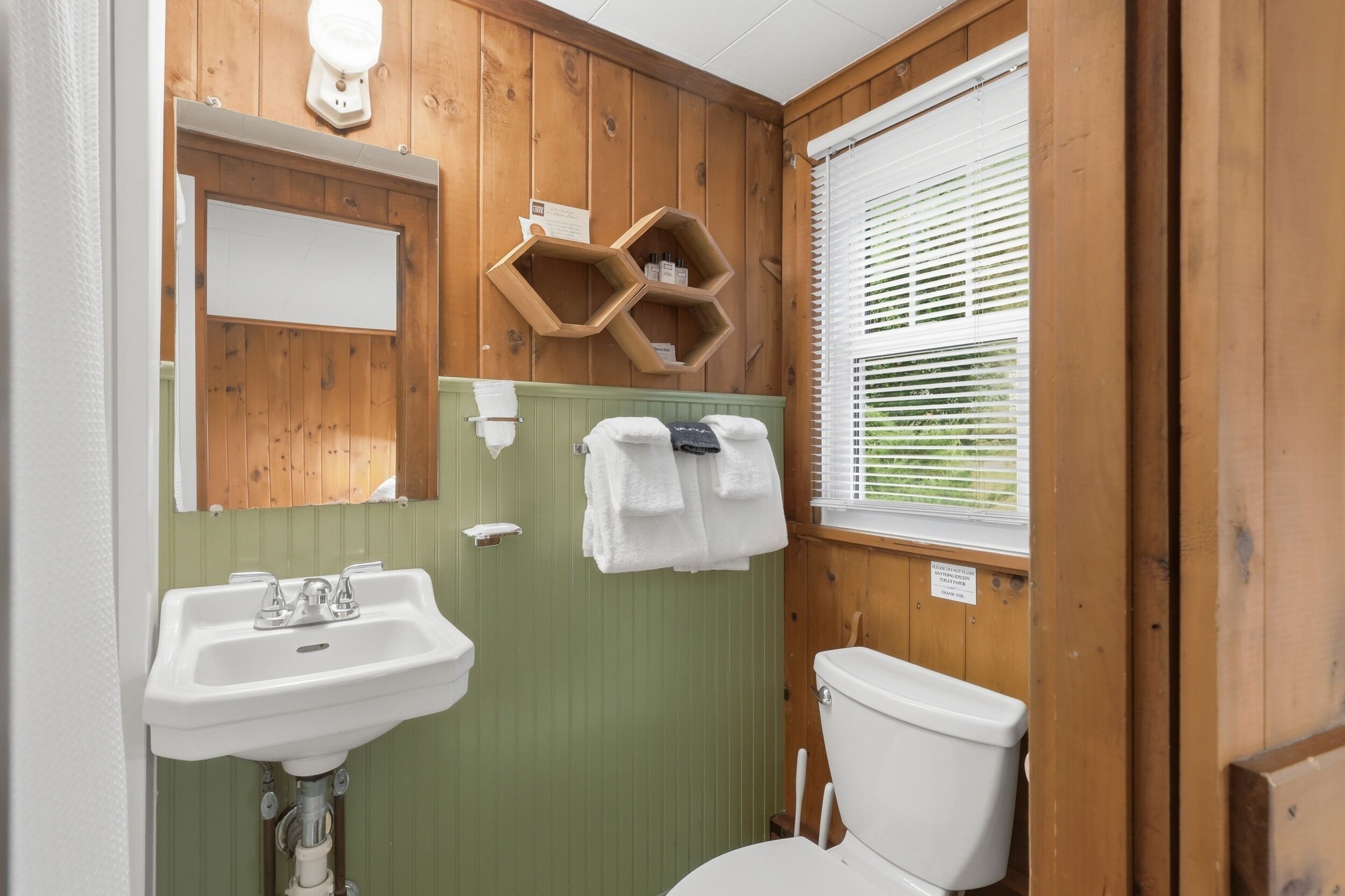 A bathroom with wooden walls, a green vanity, a white sink, a toilet, and a window with blinds.