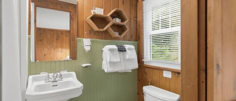 A bathroom with wooden walls, a green vanity, a white sink, a toilet, and a window with blinds.