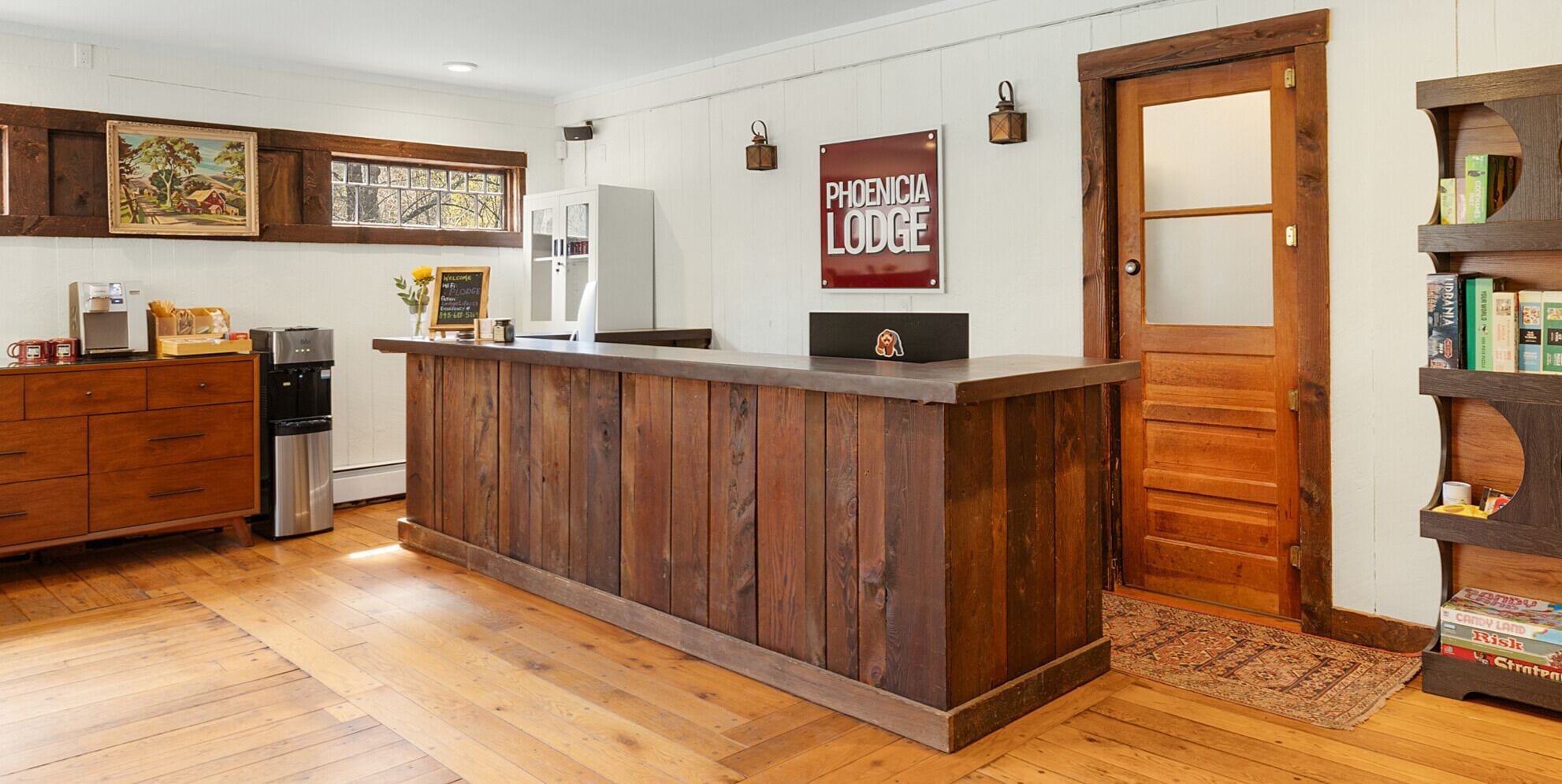 A wooden reception desk with a sign reading "PHOENICIA LODGE" in a room with wooden flooring, a coffee machine, and a bookshelf with books.
