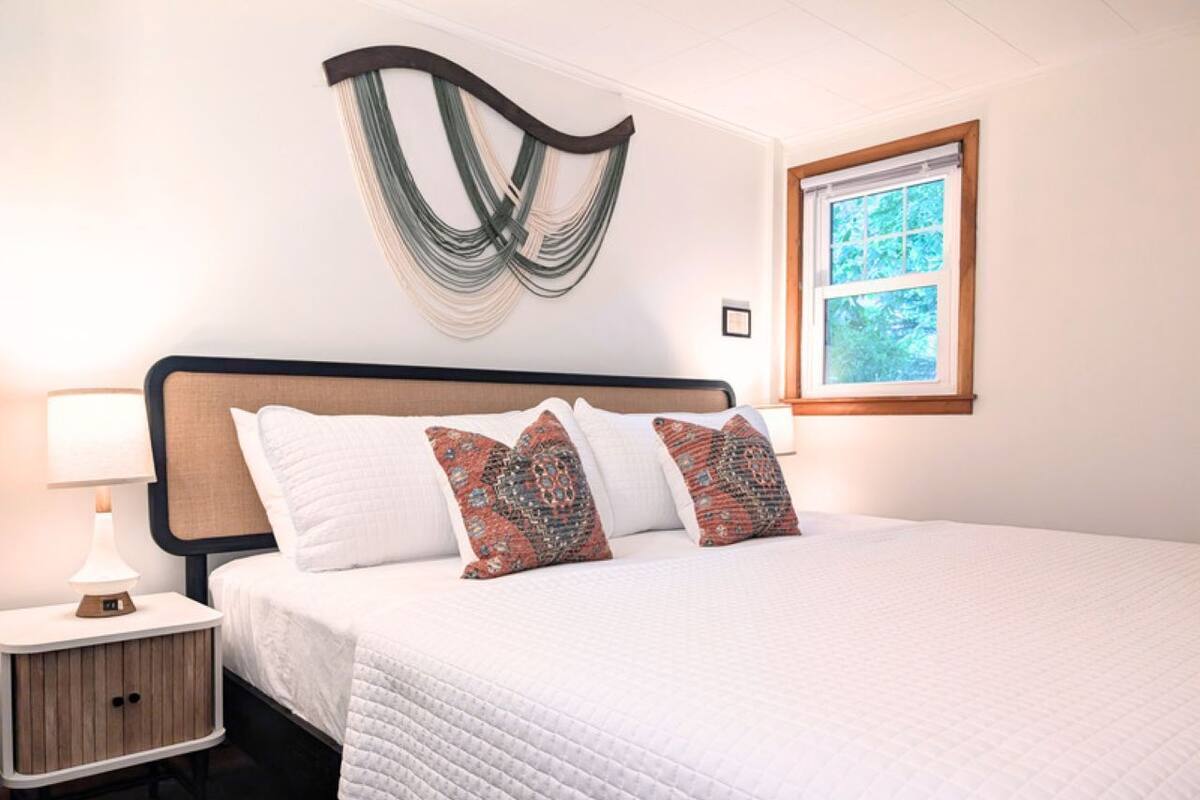 A neatly made bed with white bedding and patterned pillows, a bedside table with a lamp, and a wall-mounted abstract art piece.