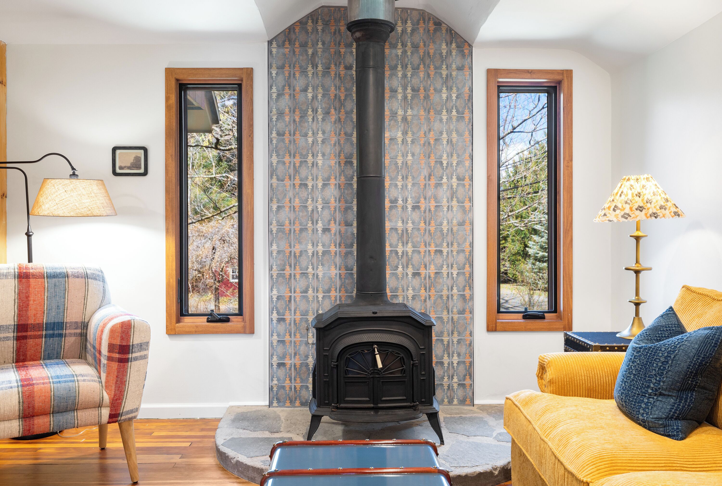 A cozy living room with a central wood-burning stove, a plaid sofa, a yellow armchair, and a patterned wall.