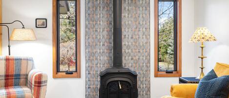 A cozy living room with a central wood-burning stove, a plaid sofa, a yellow armchair, and a patterned wall.