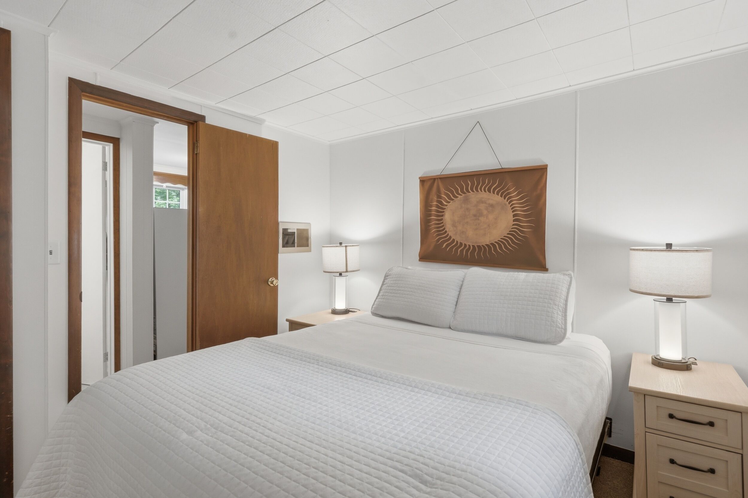 A hotel room with a bed, bedside lamps, a wooden door, and a wall-mounted sun decoration.