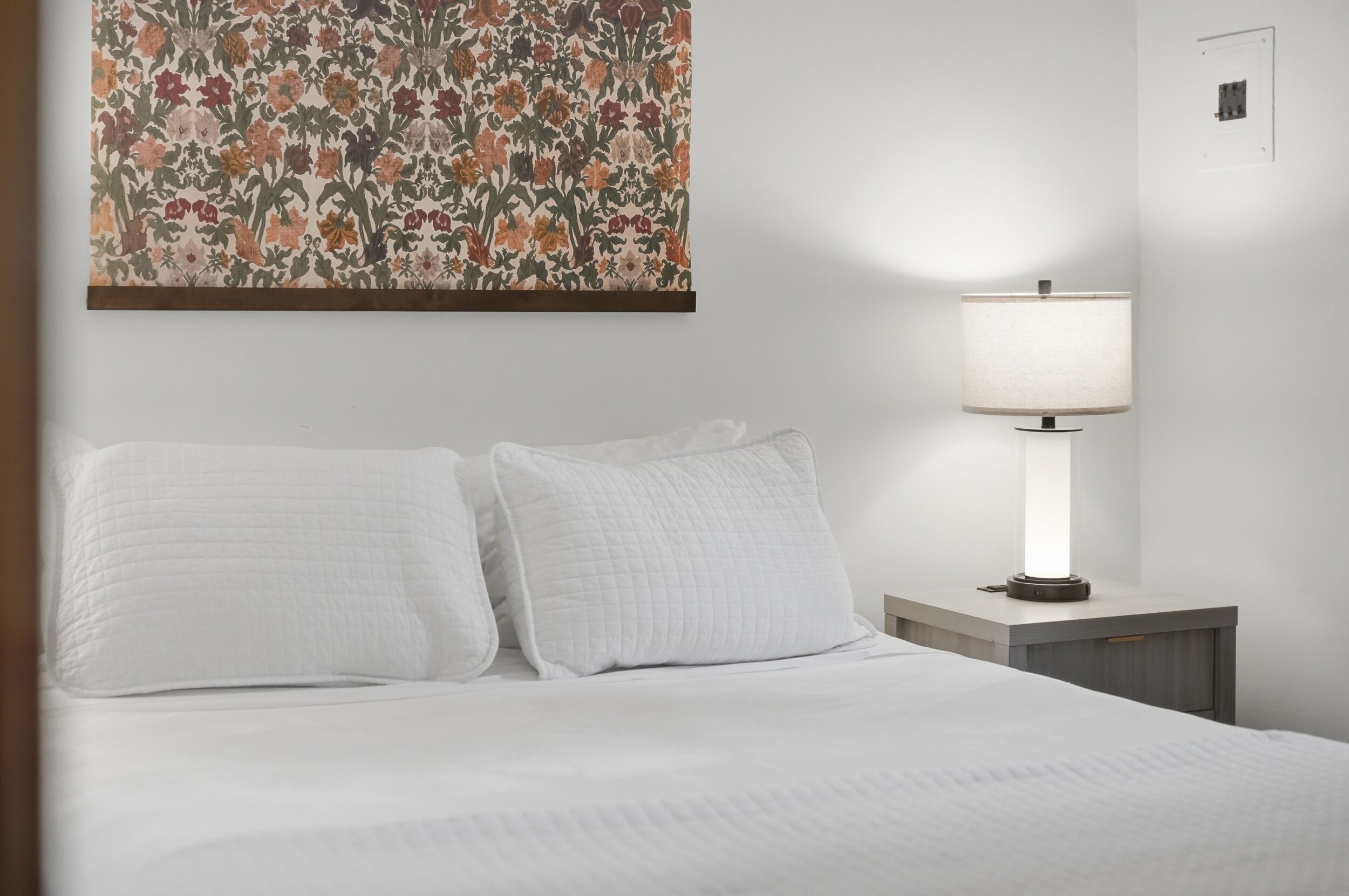 Een hotelkamer met een bed, kussens, een nachtkastje, een lamp en een bloemistisch kunstwerk aan de muur.