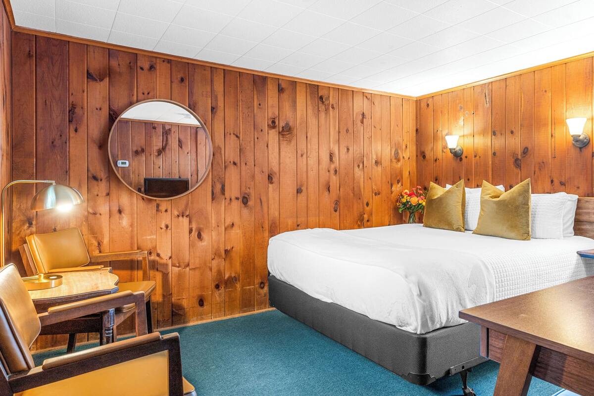 A room with a wooden paneled wall, a round mirror, a bed with white bedding, a desk with a chair, and a blue carpet.