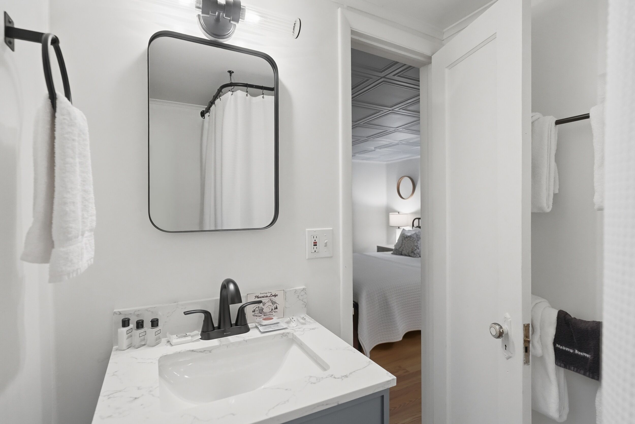 A modern bathroom with a white sink, black faucet, and a mirror above it. There are towels hanging on the wall and a shower visible in the reflection.