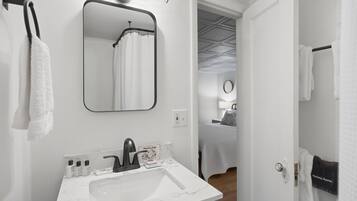 A modern bathroom with a white sink, black faucet, and a mirror above it. There are towels hanging on the wall and a shower visible in the reflection.