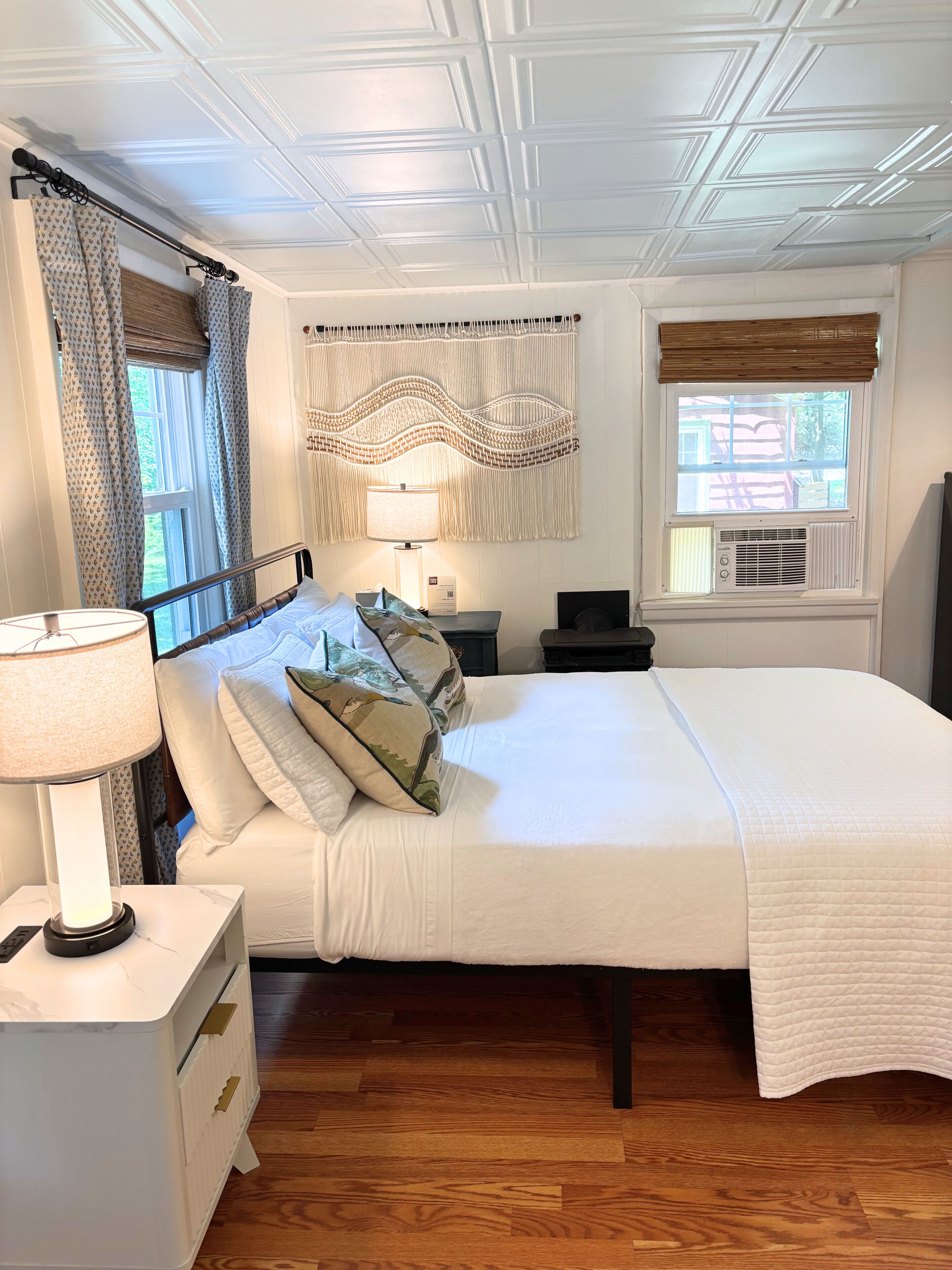 Un dormitorio con cama, mesitas de noche, una lámpara, una ventana con cortinas y un macramé colgado en la pared.