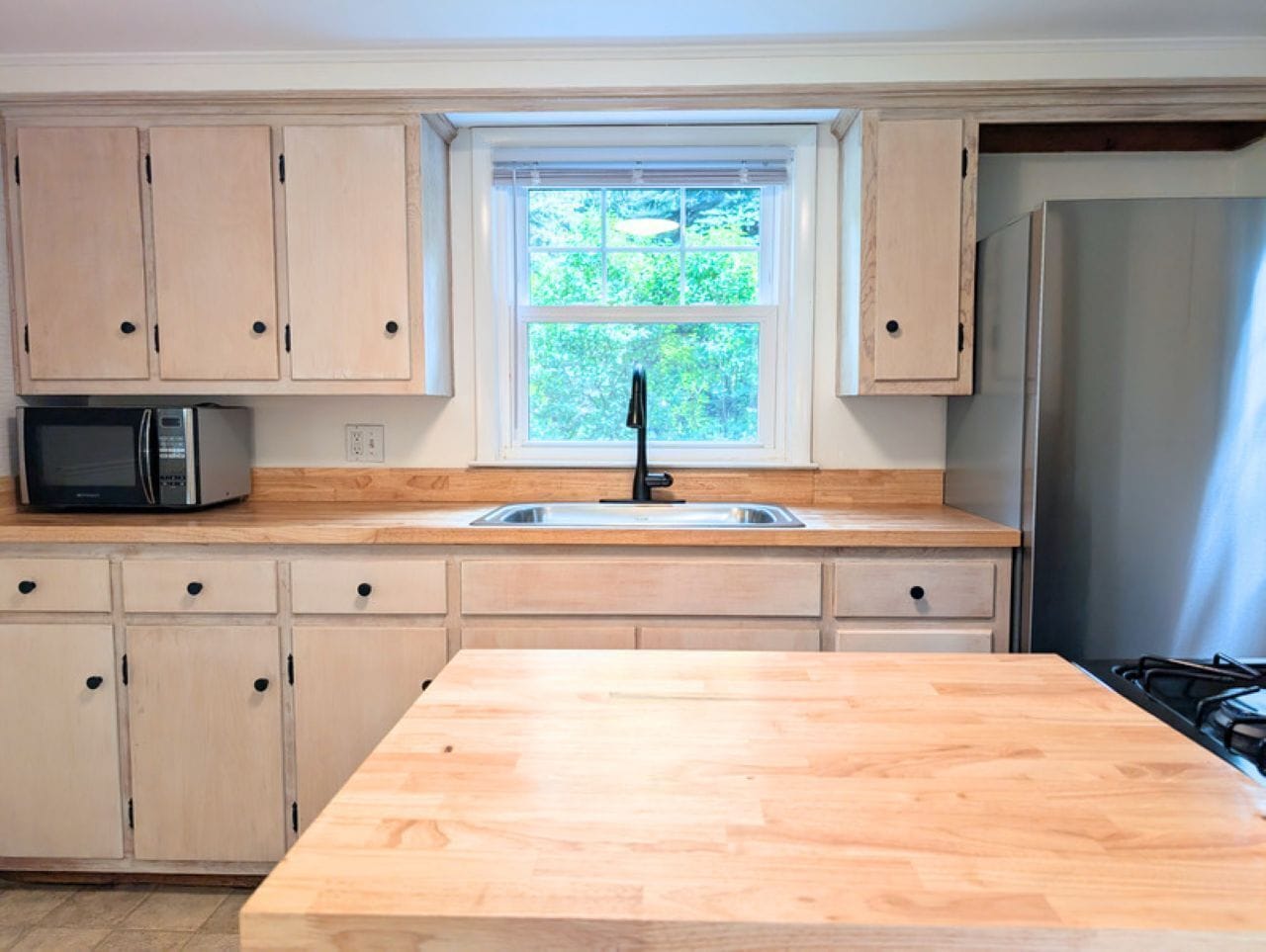 A kitchen with wooden cabinets, a microwave, a sink, and a window with a view of greenery.