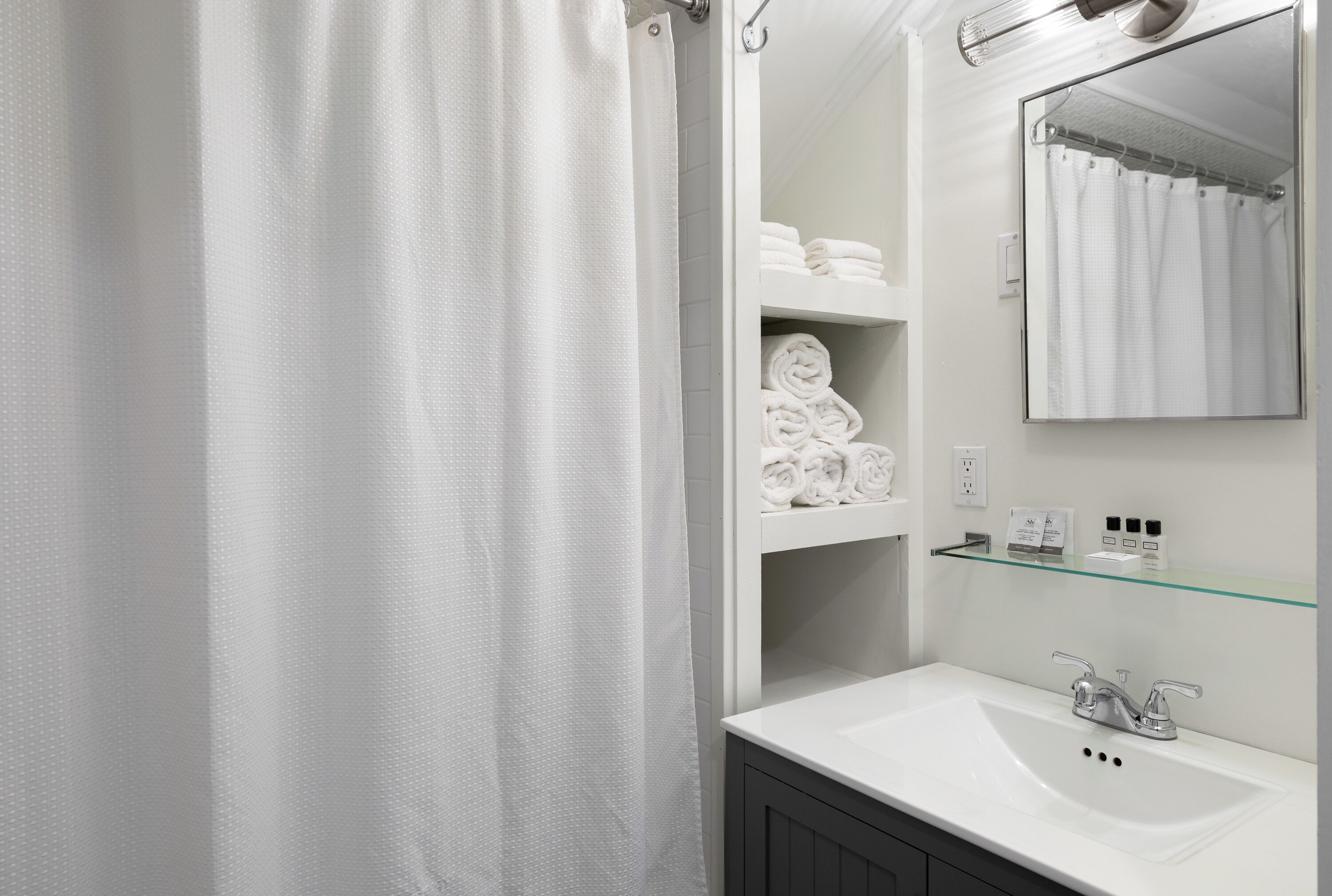 Une salle de bain avec un lavabo blanc, un miroir et des étagères où sont pliées des serviettes.