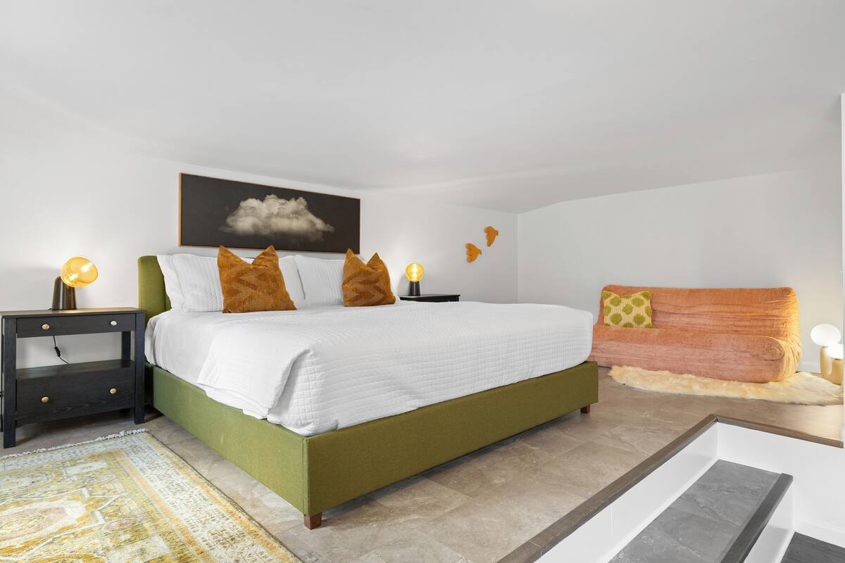 A bedroom with a green bed, white bedding, a dark nightstand, a small table with a lamp, and a sofa with orange cushions.