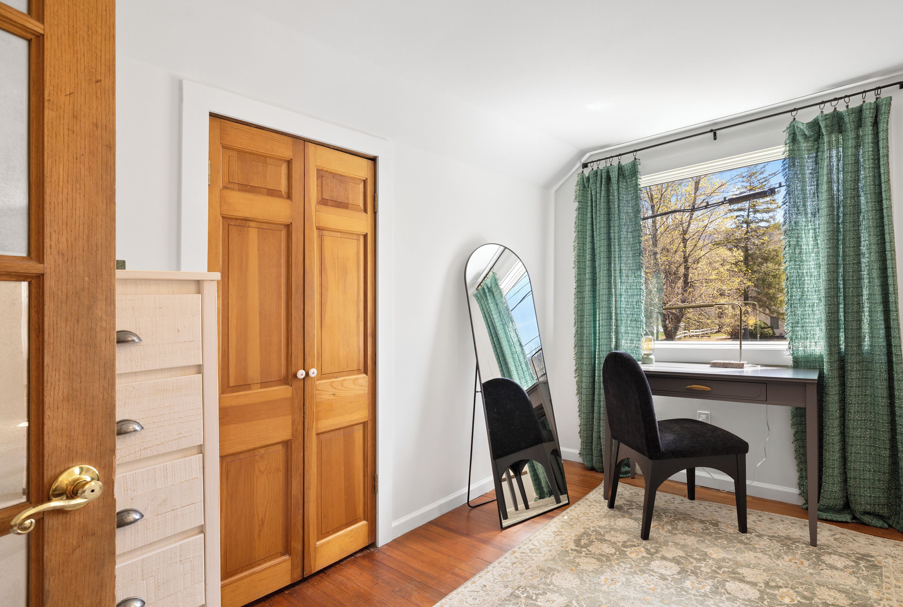 A room with a wooden wardrobe, a desk with a chair, a mirror, and a window with curtains.
