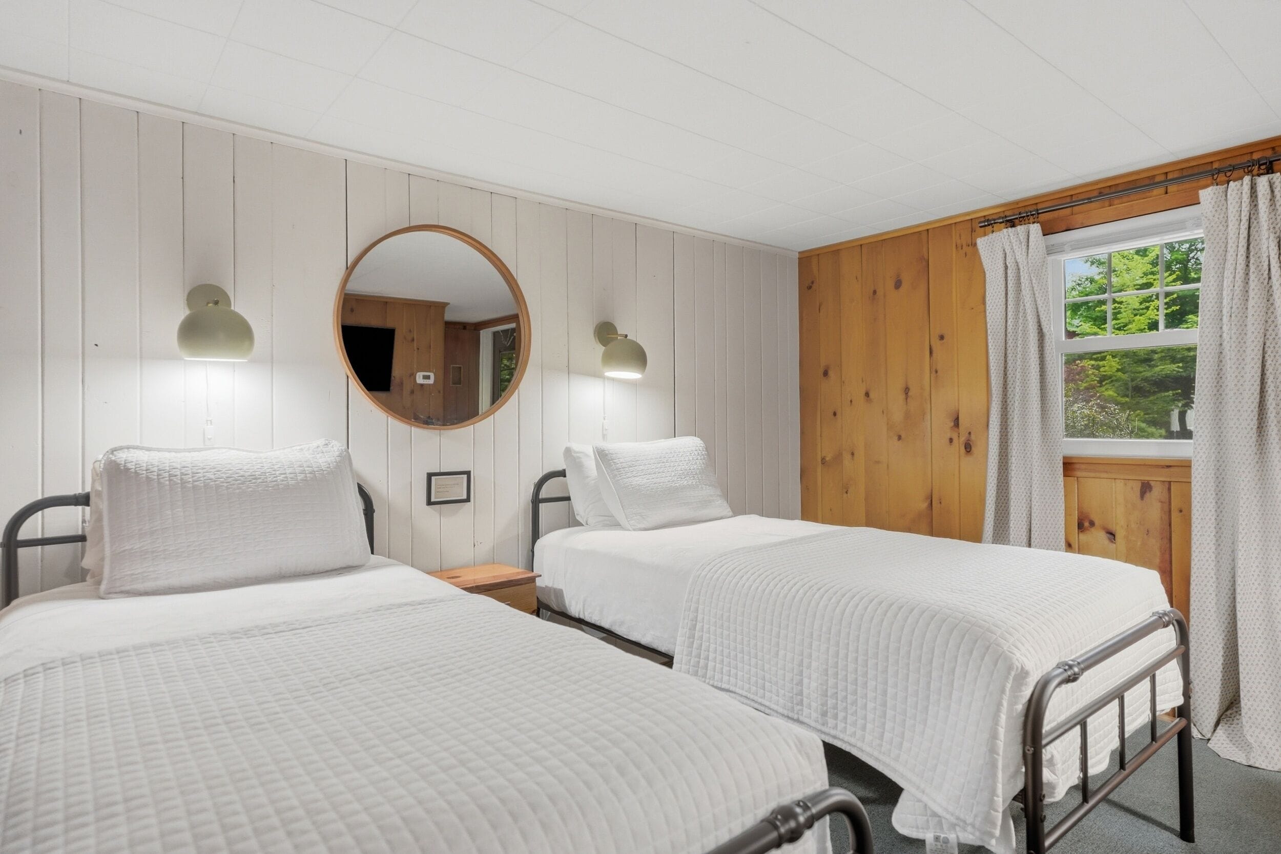 A room with two single beds, a wooden wall, a window with curtains, and a round mirror.