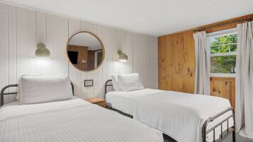A room with two single beds, a wooden wall, a window with curtains, and a round mirror.