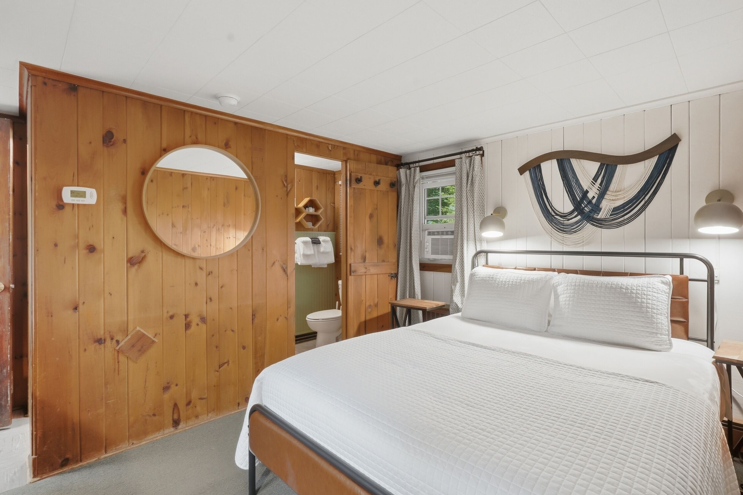 Un dormitorio con puerta de madera, espejo redondo, cama con ropa blanca, hamaca fijada a la pared y una mesita.