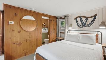Un dormitorio con puerta de madera, espejo redondo, cama con ropa blanca, hamaca fijada a la pared y una mesita.