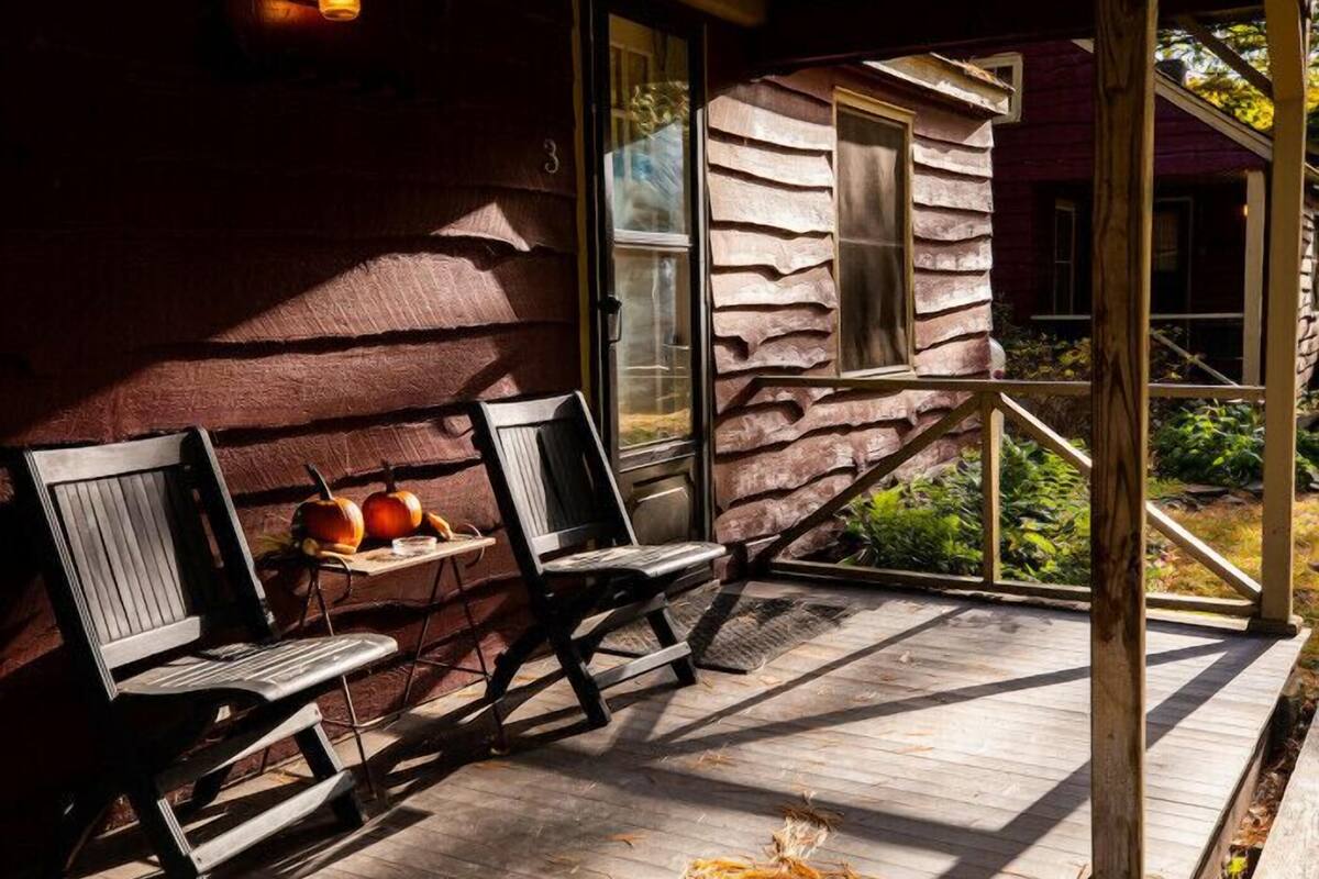 A wooden porch with two chairs and a table, a pumpkin, and a house with a unique stone facade.