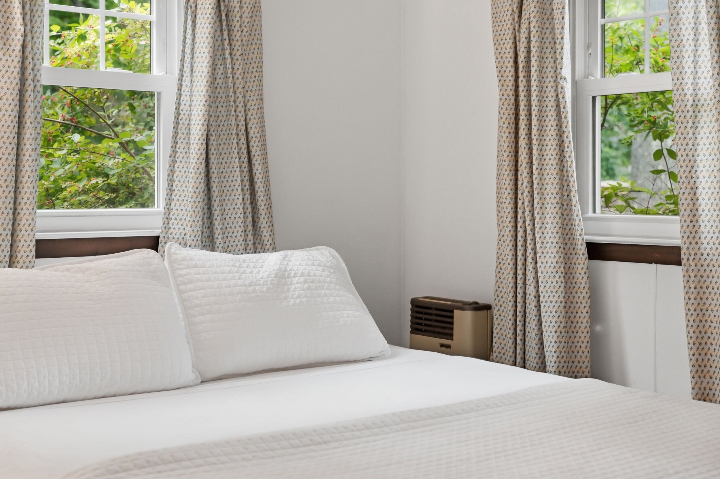 A bedroom with a bed, white bedding, a bedside table, a heater, and curtains with a leaf pattern.