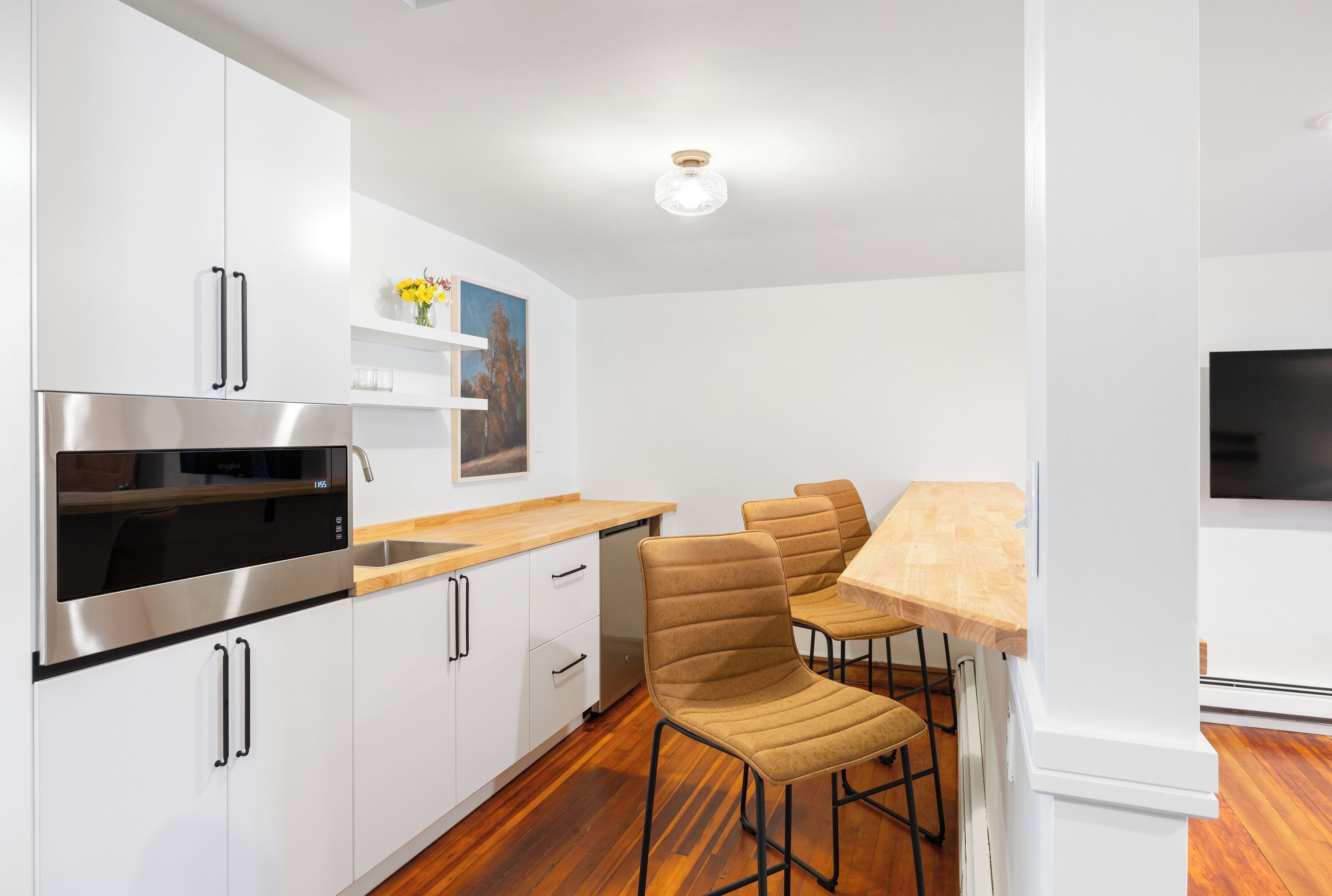 Una cocina moderna con armarios blancos, electrodomésticos de acero inoxidable y una encimera de madera. Hay dos taburetes de bar y una mesita con tapa de madera clara.