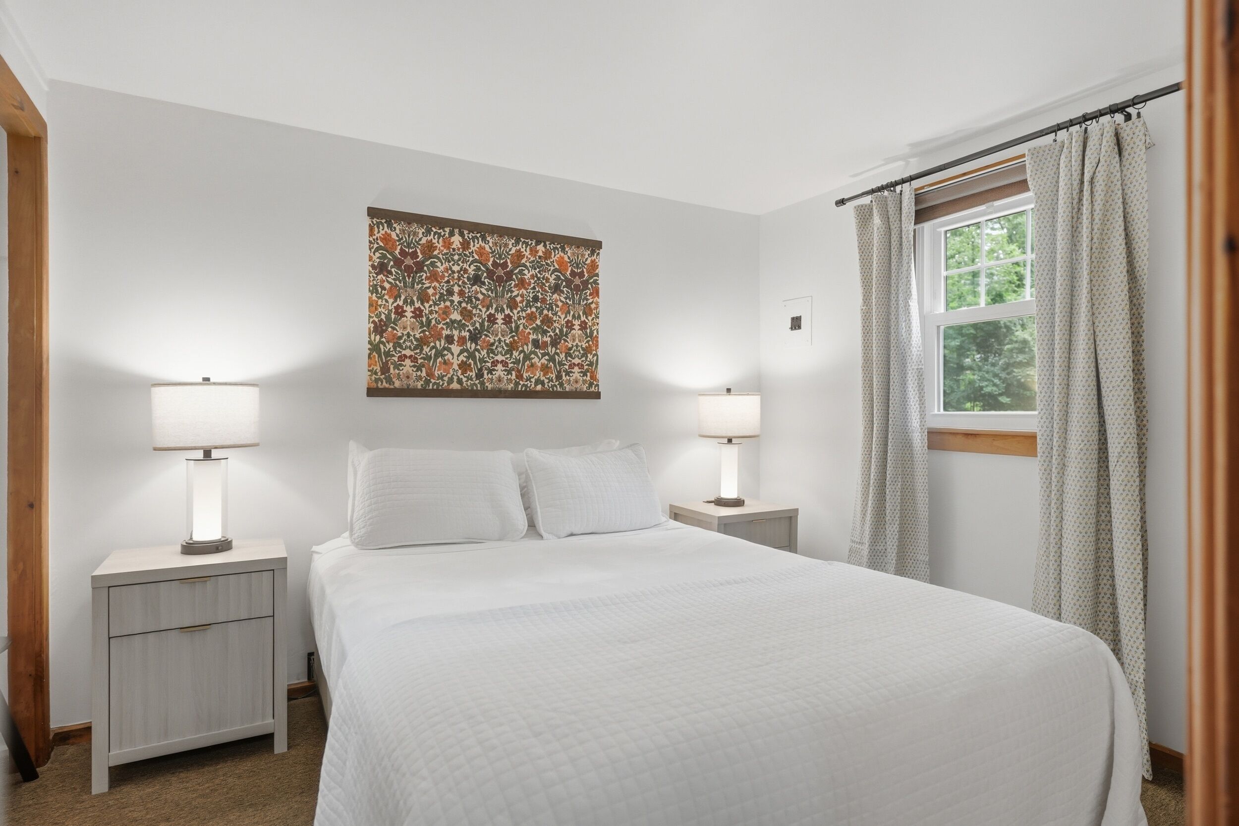 A bedroom with a large bed, two bedside tables with lamps, a floral artwork on the wall, and a window with curtains.