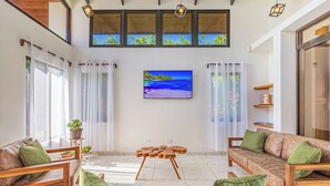 Een ruime woonkamer met een grote flatscreen televisie aan de muur, houten meubels en uitzicht op de natuur door grote ramen.