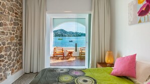 Un dormitorio con pared de piedra, cama con manta colorida, mesita de noche con lámpara y vistas a un balcón con mesa y sillas que dan a un cuerpo de agua.