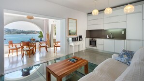 Una sala de estar moderna con una mesa de centro de vidrio, un sofá con un cojín estampado, y un comedor con sillas y mesa de madera. Se ve una cocina con armarios blancos y vistas al mar a través de un arco.