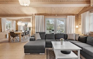 A cozy cabin interior with a sectional sofa, dining table, chairs, and large windows offering views of the outdoors. - Awesome home in Væggerløse with sauna (Væggerløse)