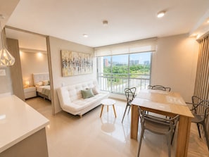 A modern living room with a white sofa, a dining table, and chairs. There is a large window offering a view of greenery and buildings. - Apartamentos Palmar - by SOHO (Puerto Colombia)