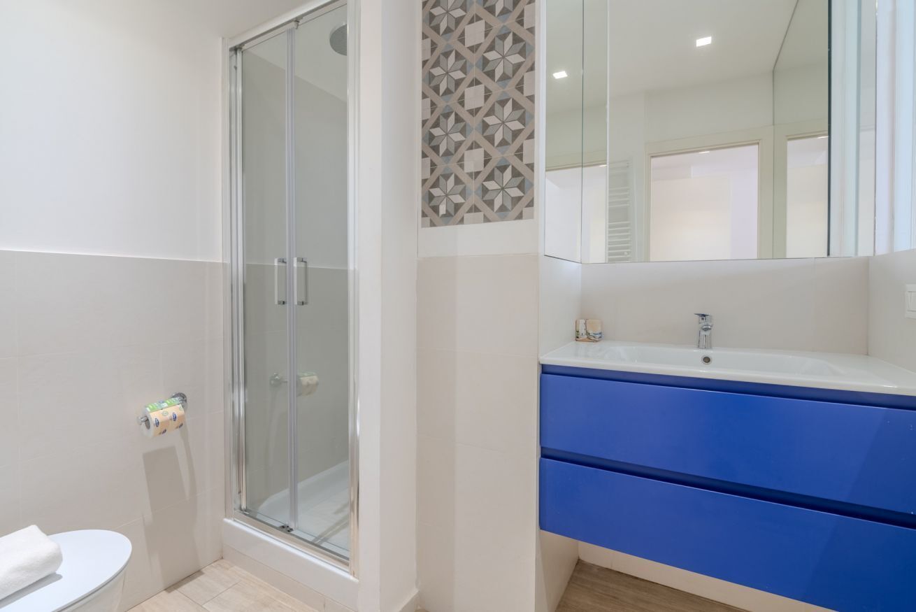 Une salle de bain avec une douche à parois de verre, des toilettes blanches, un lavabo avec des meubles bleus et un panneau décoratif mural.