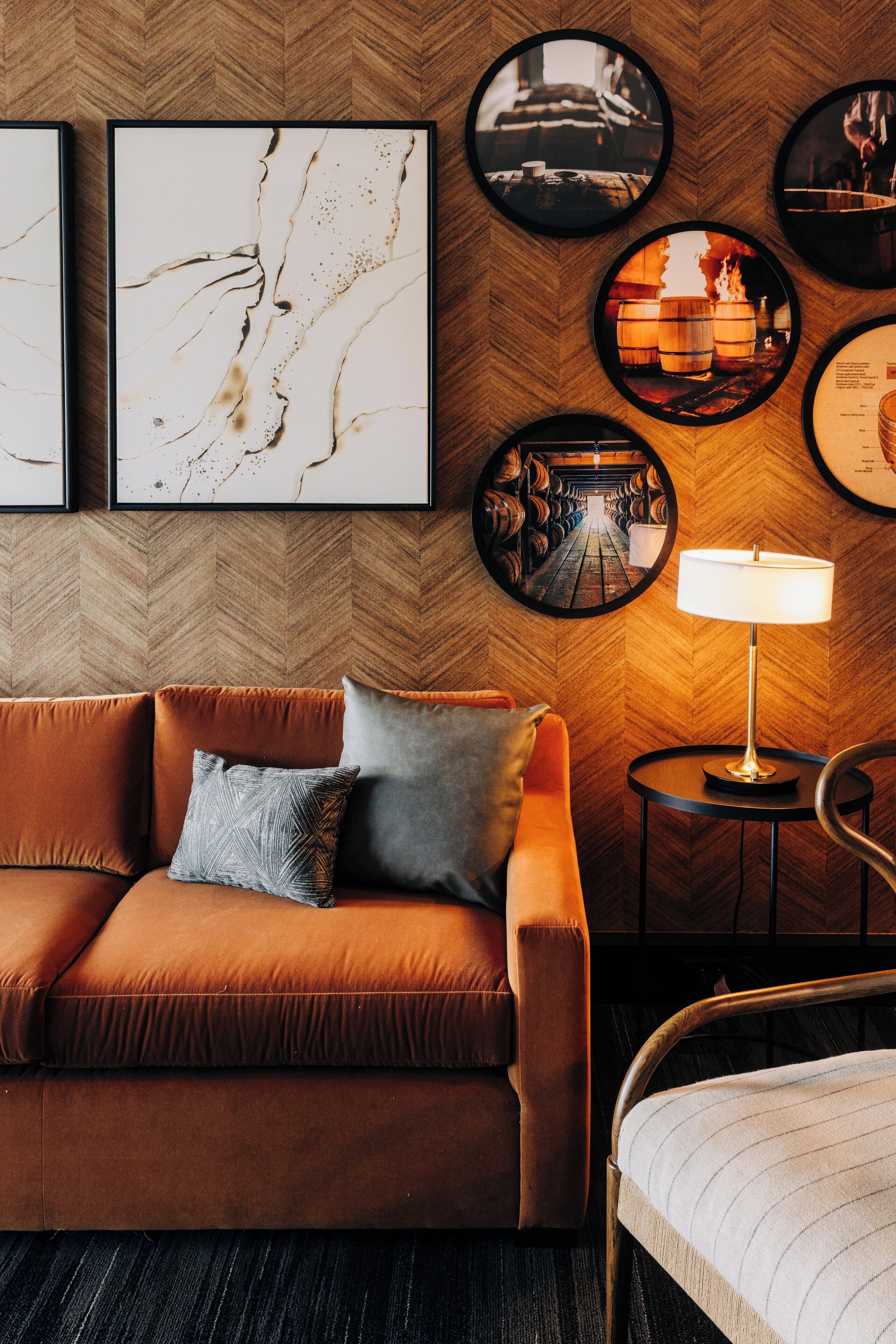 A modern hotel room with a herringbone-patterned wooden wall, a brown leather sofa, a round side table with a lamp, and framed artwork.