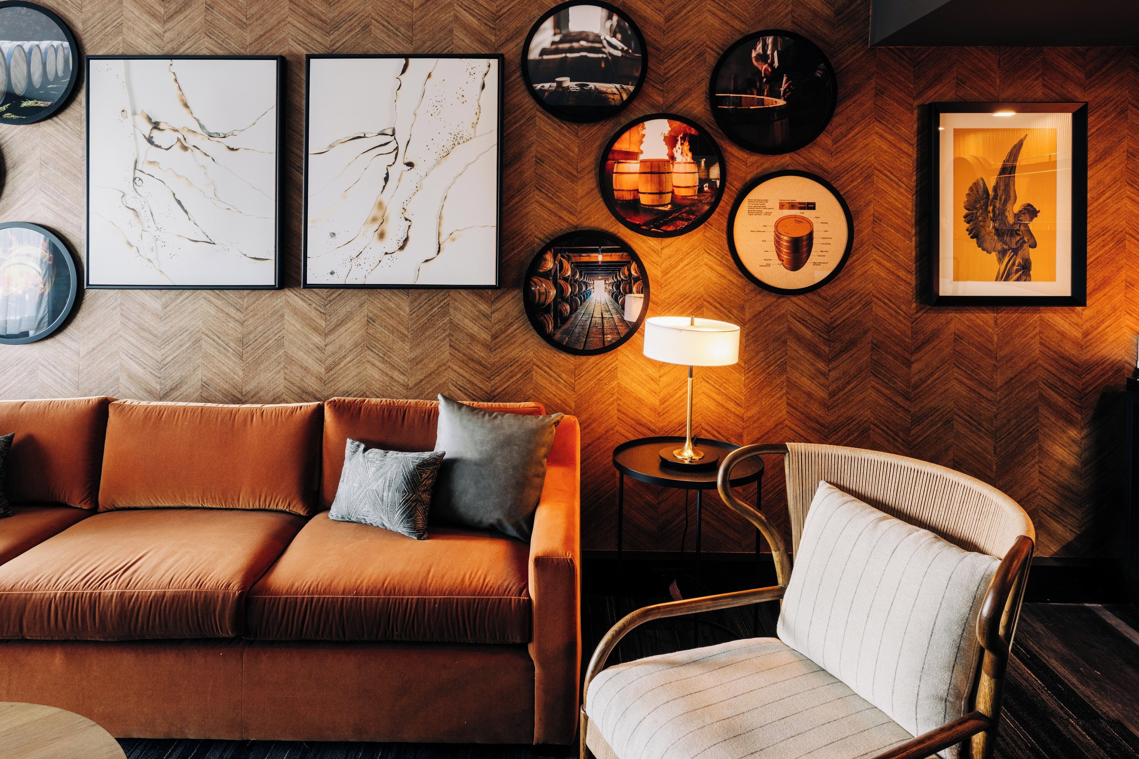 A modern living room with a herringbone-patterned wooden wall, a brown sofa, a striped armchair, and various framed artworks.