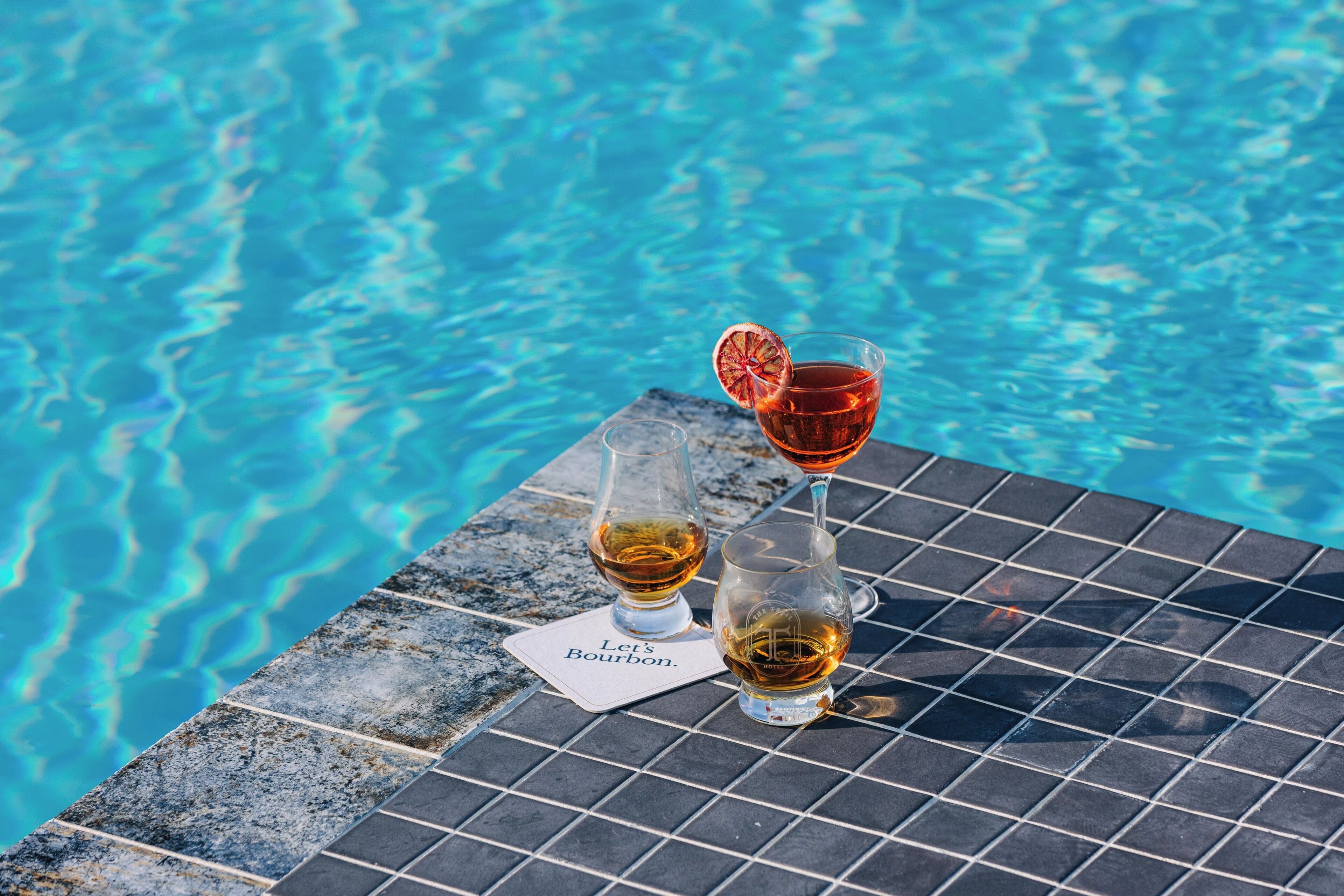Deux verres de whisky sur une table carrelée au bord de la piscine, avec une pancarte sur laquelle on peut lire : « À la vôtre, avec du bourbon ! »