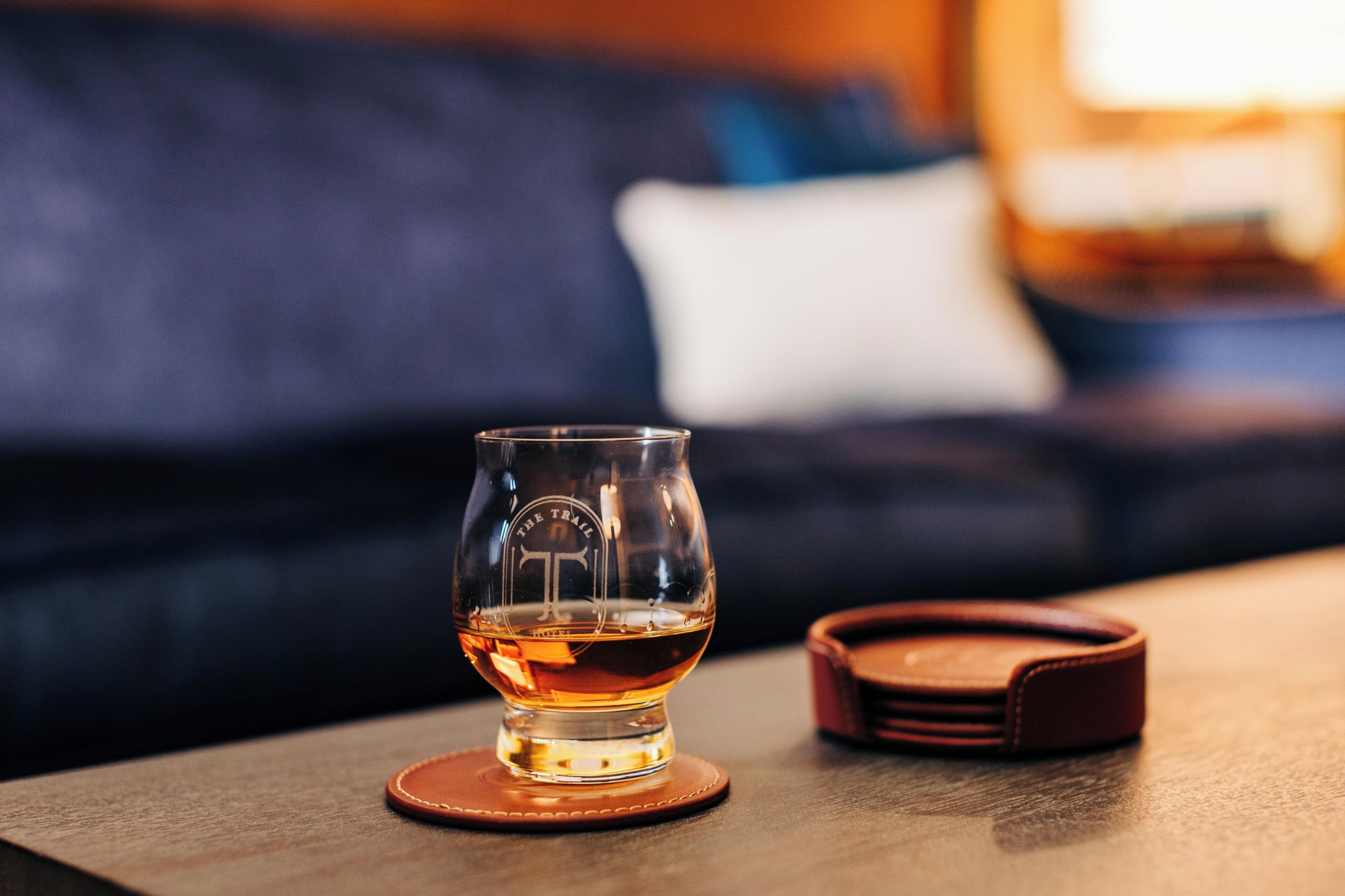 A clear glass of whiskey on a coaster with a leather case beside it on a wooden table.