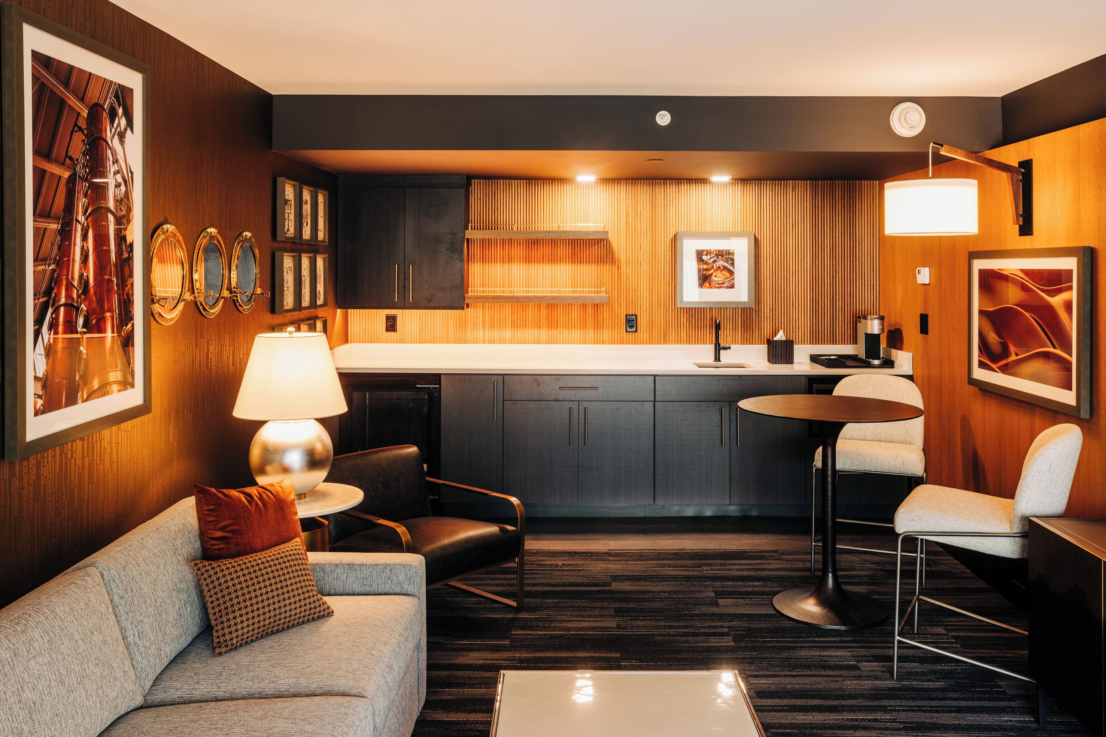 A modern hotel room with a sofa, a dining table, a kitchen area, and a bar.
