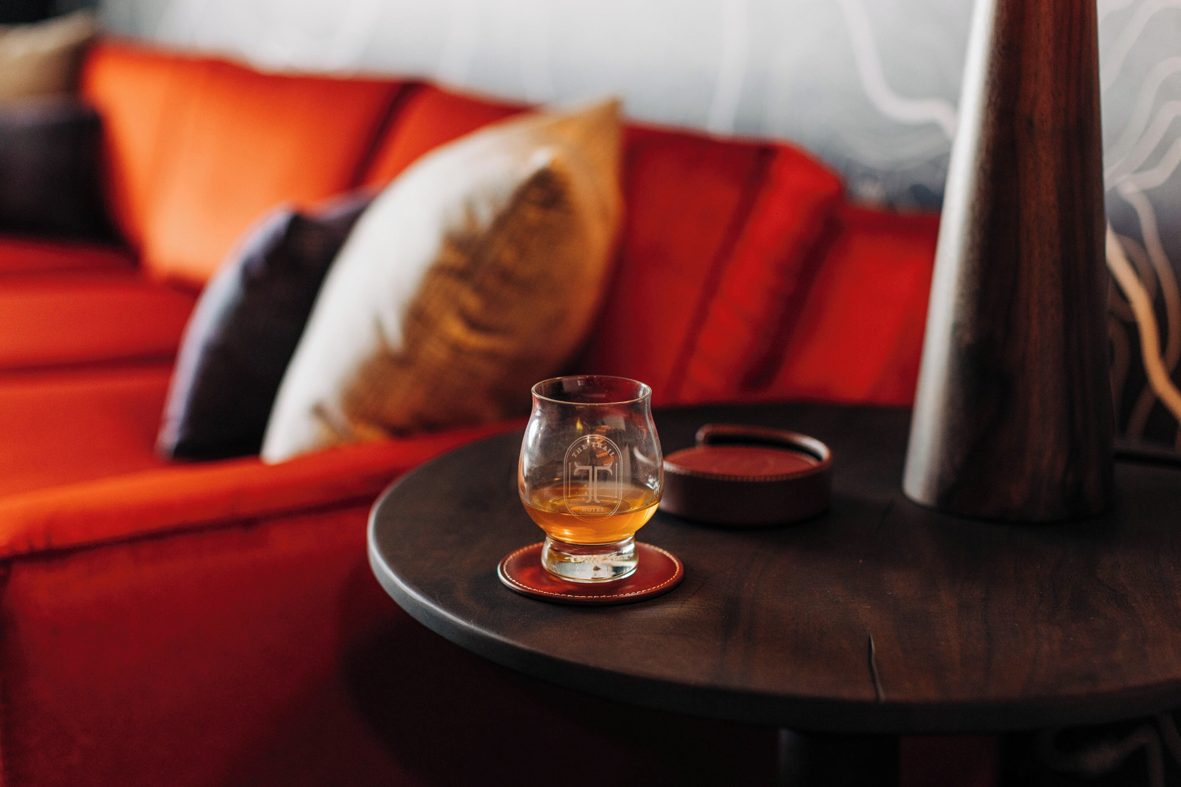 Un vaso de líquido ámbar sobre un posavasos, un tazón pequeño y un recipiente metálico sobre una mesa redonda en una habitación con muebles tapizados en rojo y un papel tapiz estampado.