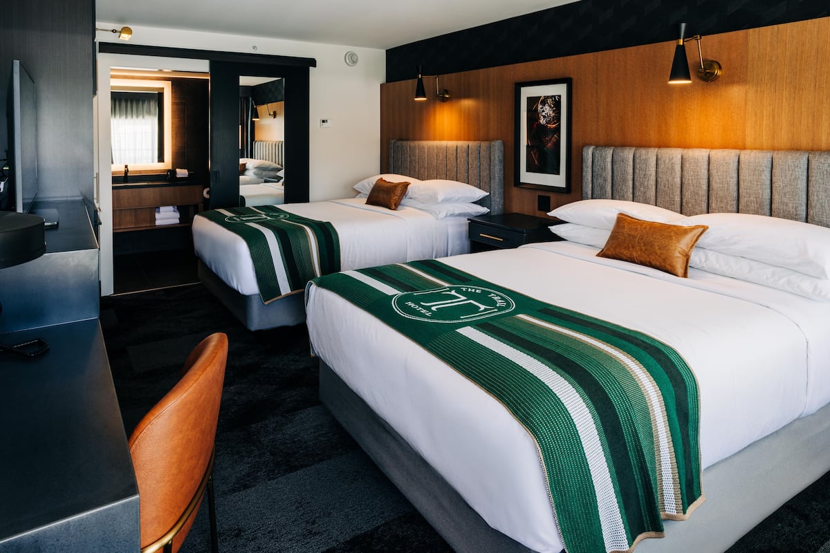 A hotel room with two beds, a desk, and a chair. There is a mirror, a framed picture on the wall, and a wooden headboard.