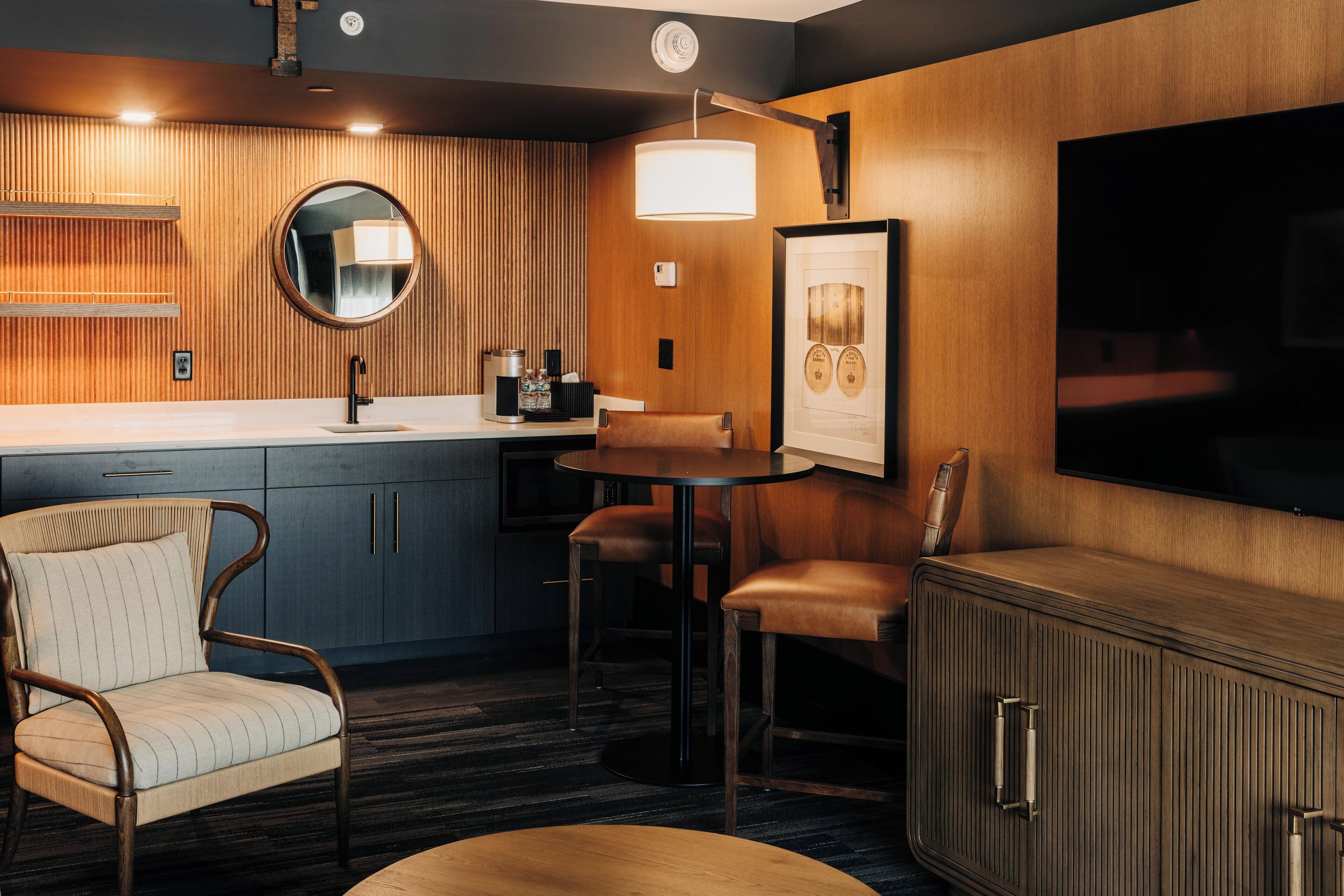 Ett modernt hotellrum med en väggpanel i trä, en rund spegel, ett litet runt bord, två läderfåtöljer, ett skåp och en platt-TV.