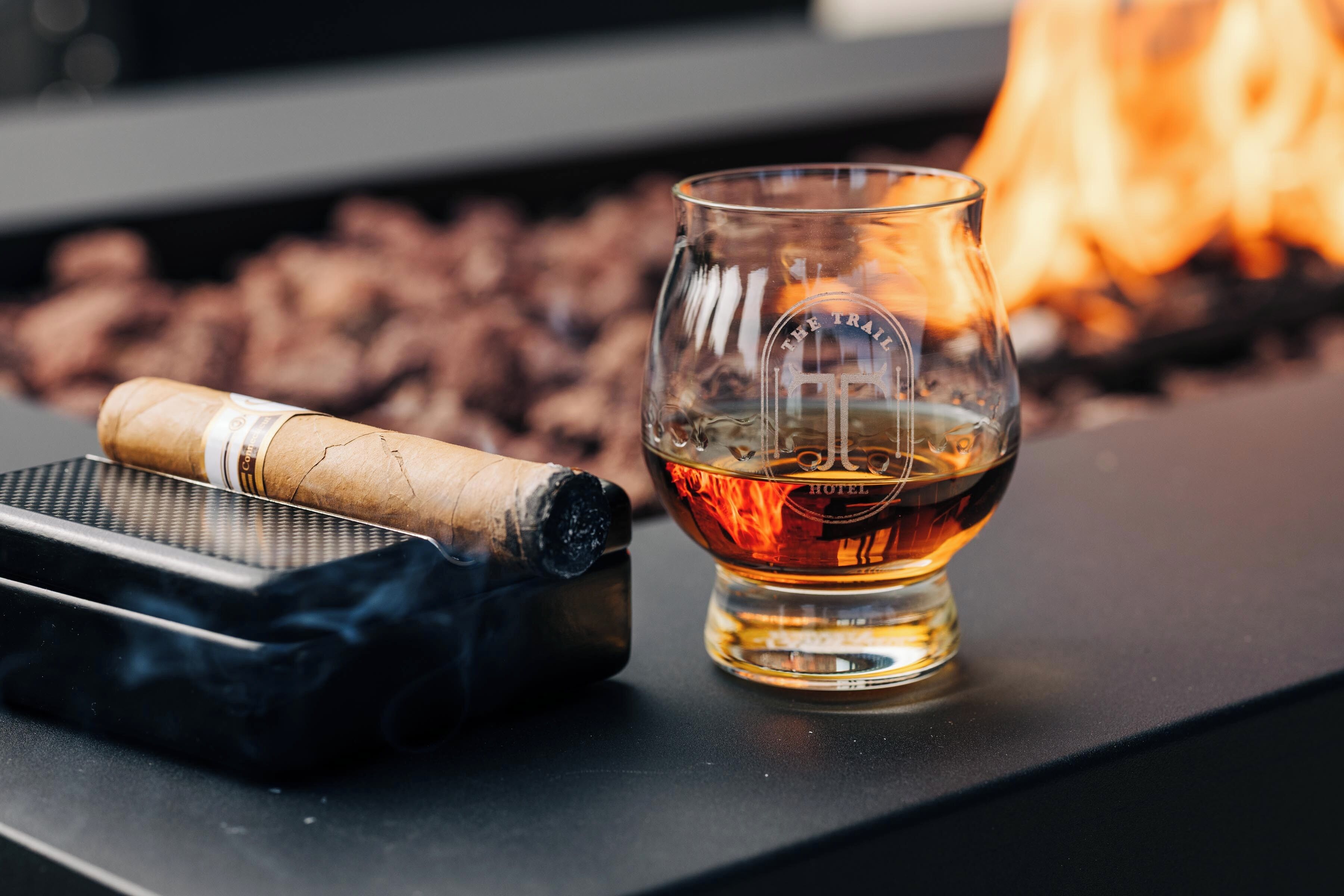 A glass of whiskey and a cigar on a table with a fire in the background.
