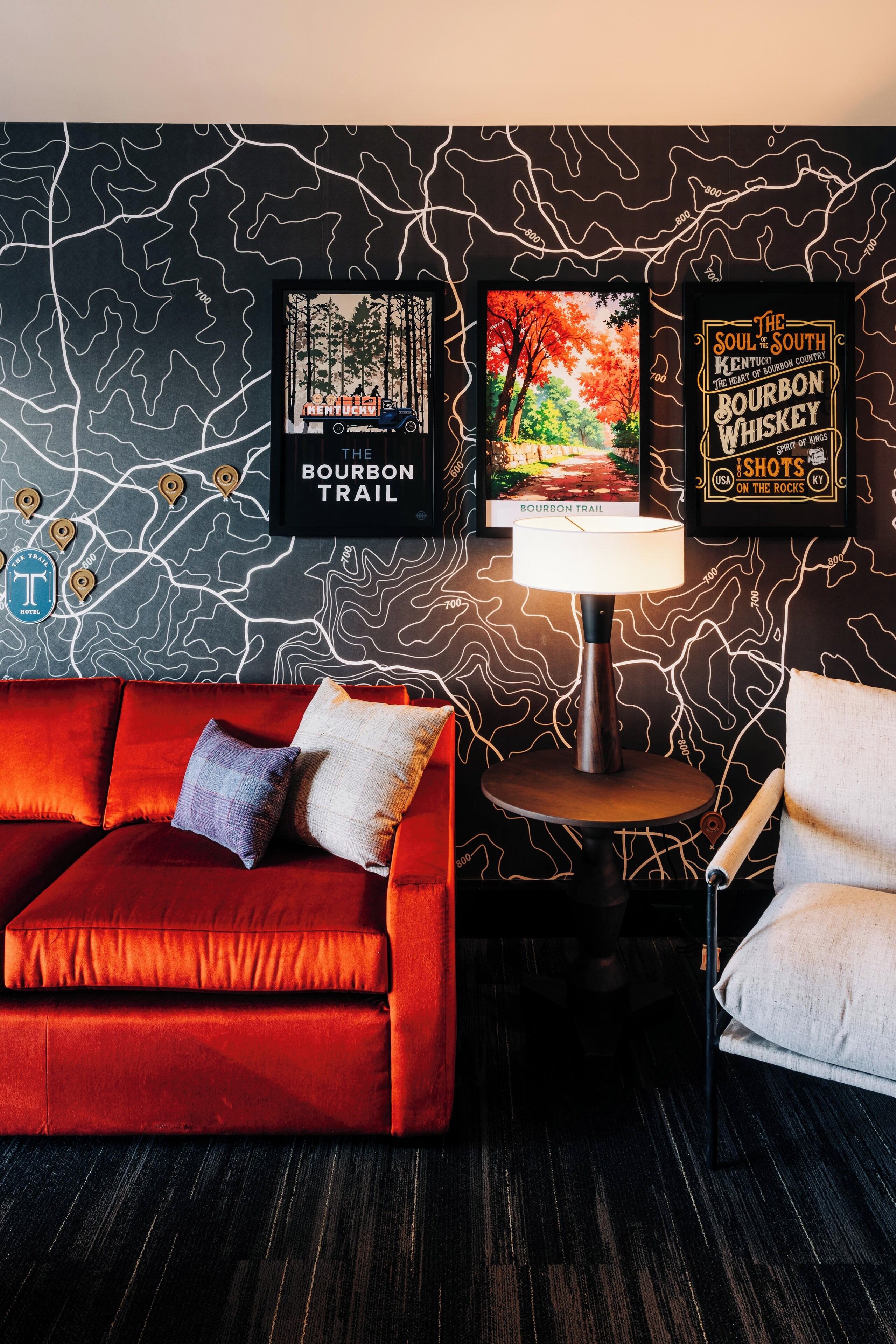 A room with a red sofa, a white armchair, a small table with a lamp, and wall art featuring bourbon trail and bourbon whiskey shots.