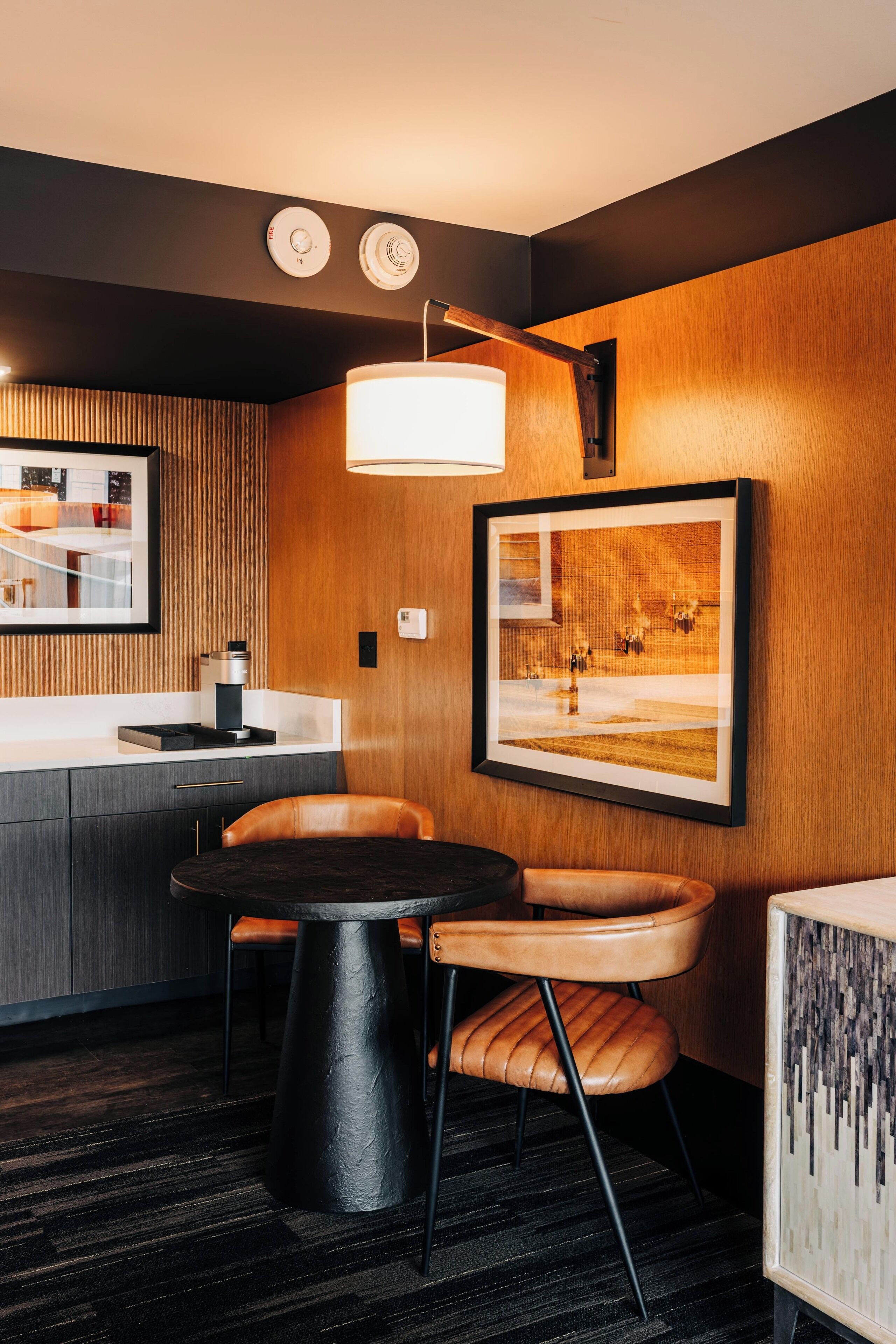 Ein modernes Hotelzimmer mit einem runden Esstisch, zwei Ledersesseln, einer Kaffeemaschine, einem gerahmten Kunstwerk und einer außergewöhnlichen Wandleuchte.