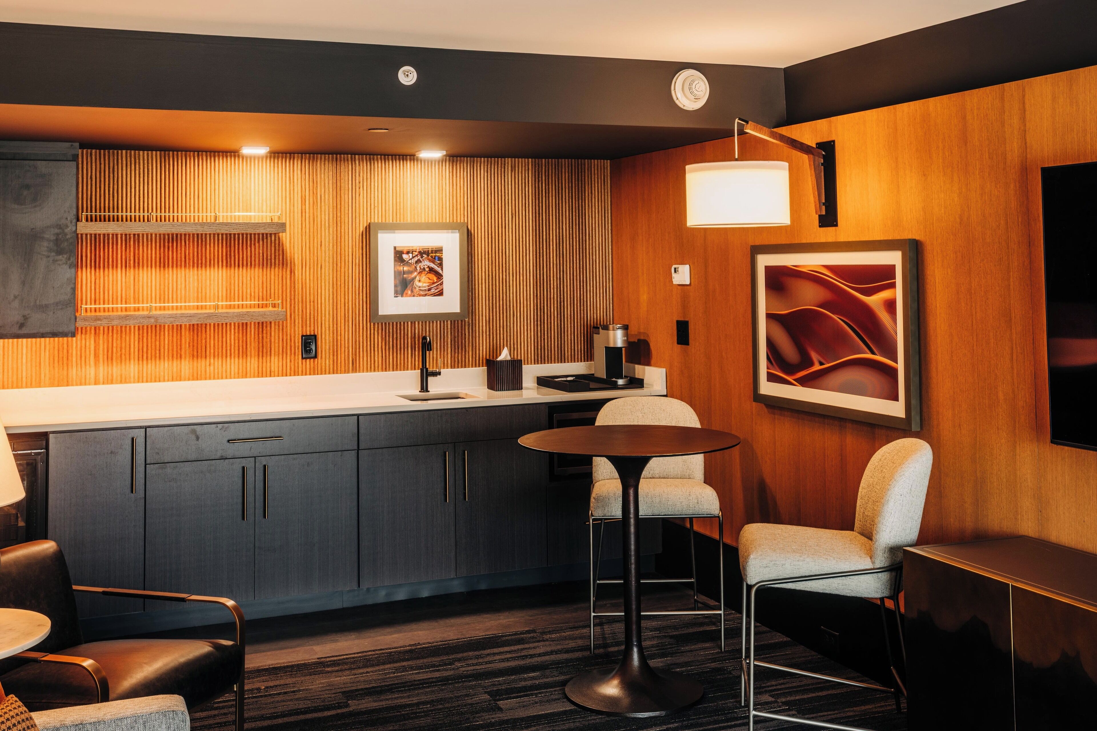 Ein modernes Hotelzimmer mit einer Wandpaneele aus Holz, einer Küchenzeile, einem runden Tisch und einer Bar.