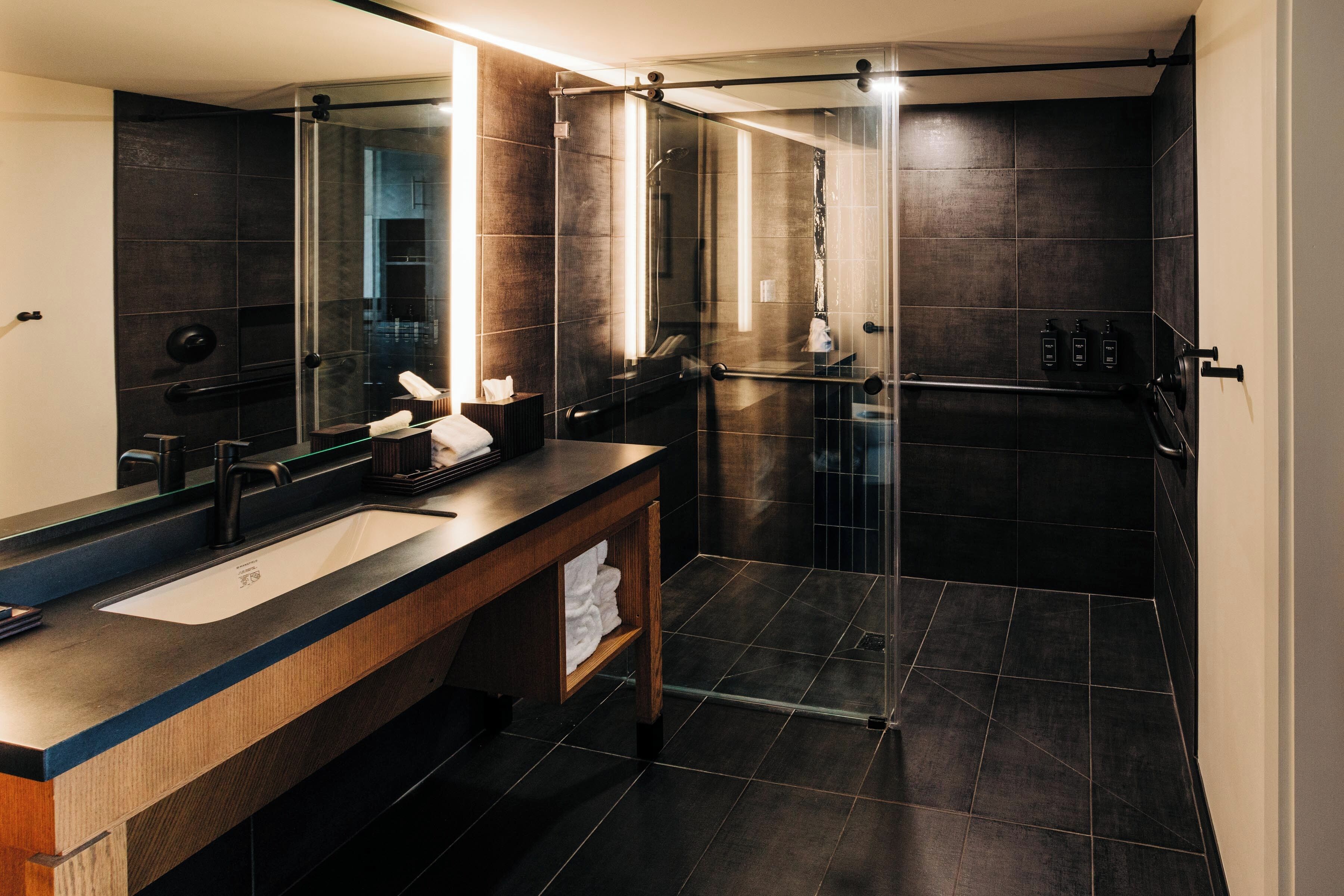 Un baño moderno con un espejo grande, baldosas oscuras, un tocador de madera y una cabina de ducha de vidrio.