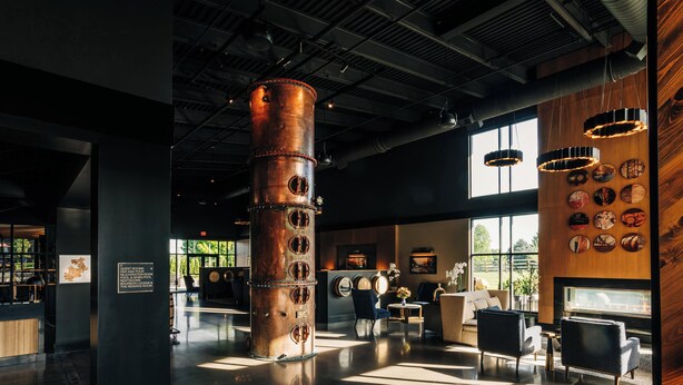 A spacious lobby with a large cylindrical column, seating areas, and a wall decorated with various items. - The Trail Hotel (Bardstown)