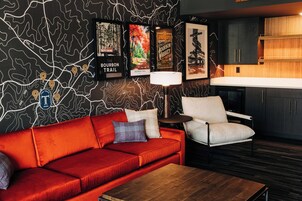 A modern hotel room with a red sofa, a white armchair, a wooden coffee table, and a map wall with Bourbon Trail markers.