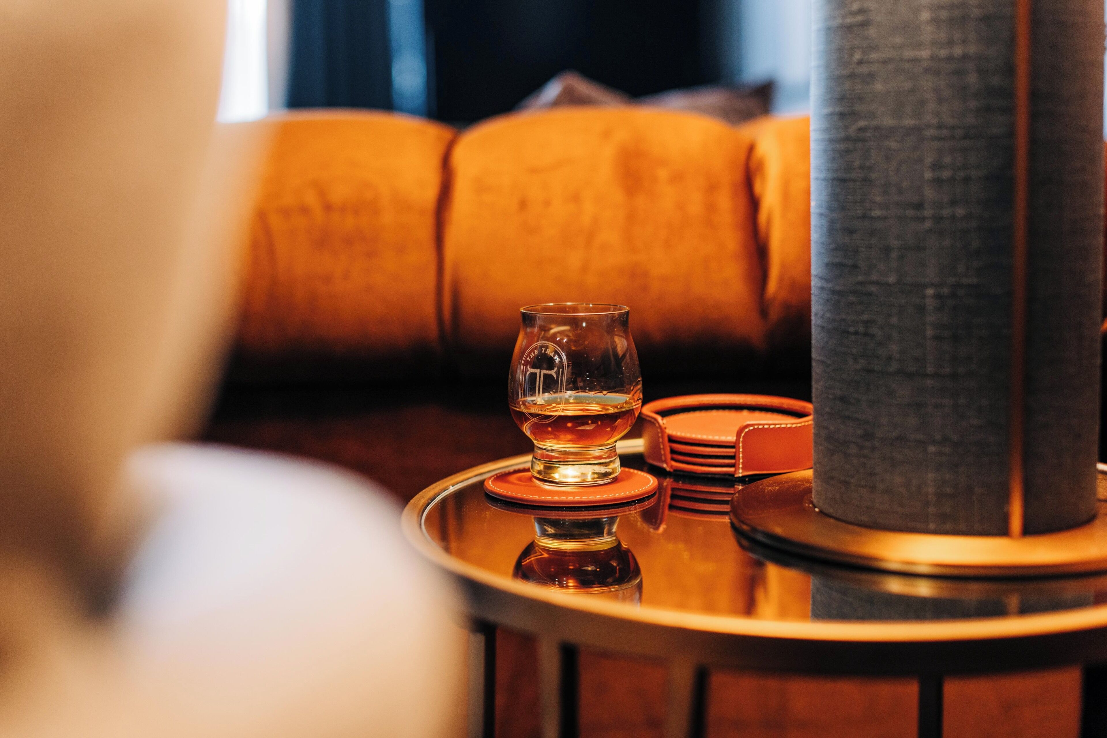 A glass of whiskey on a round table with a leather coaster, a stack of coasters, and a tall, cylindrical object, possibly a candle holder, in a warmly lit room with a sofa in the background.