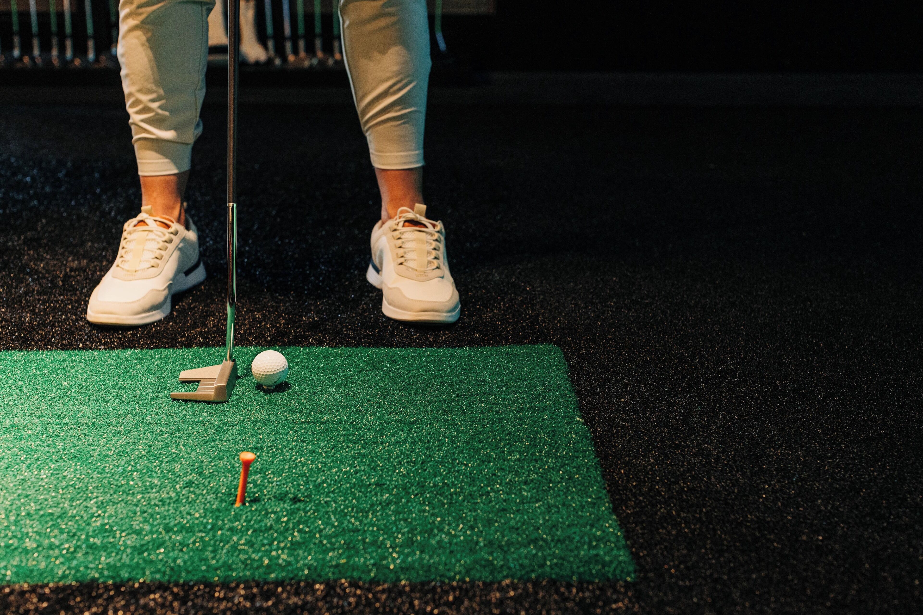 Una persona de pie sobre un tapete verde, con una pelota de golf y un elevador, preparándose para golpear.