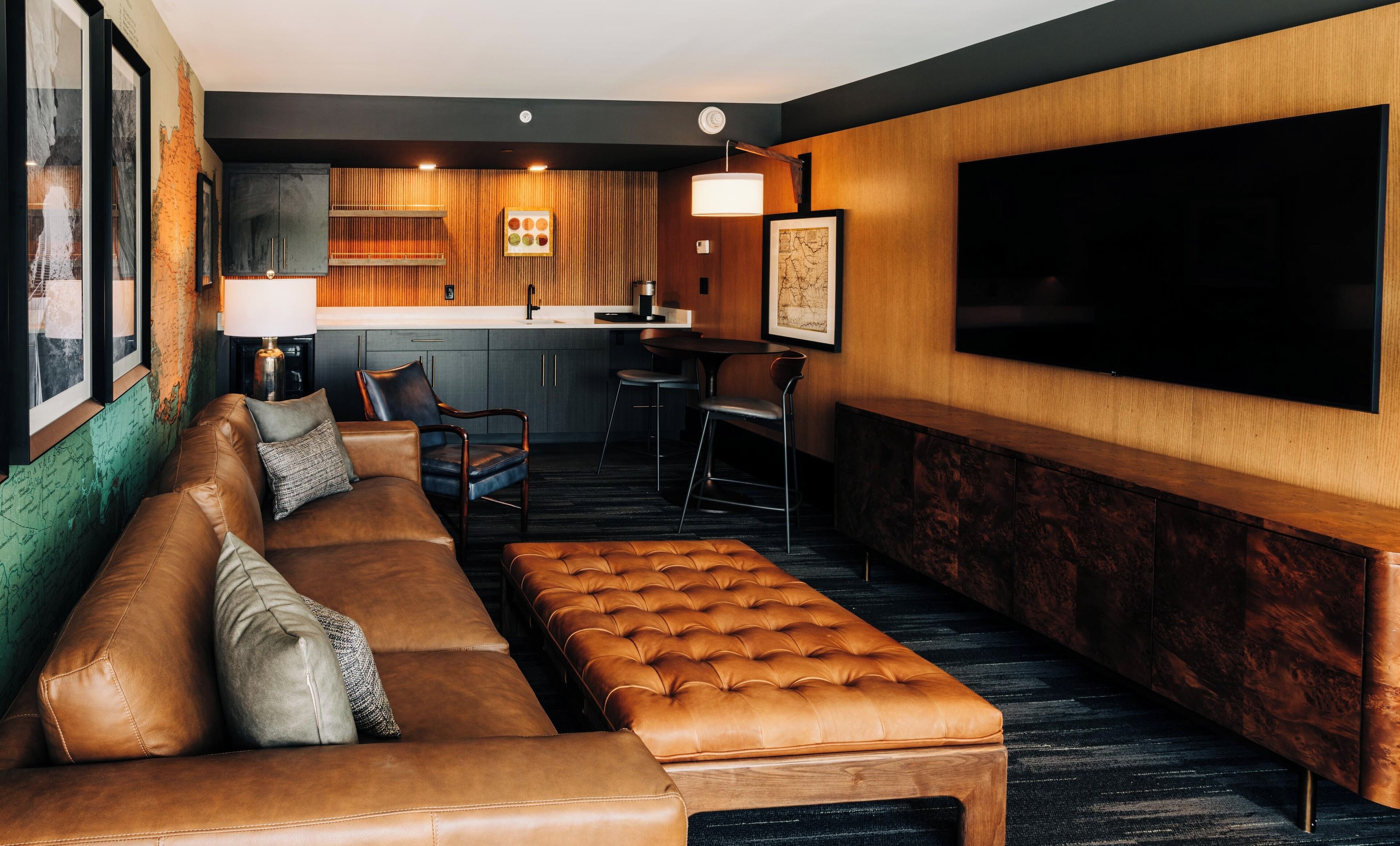 Ein moderner Lounge-Bereich mit einer Ledersofa, einem gepolsterten Hocker, einer Wandpaneele aus Holz, einem großen Flachbildfernseher und einer Bar mit Hockern und einer Holztresen.