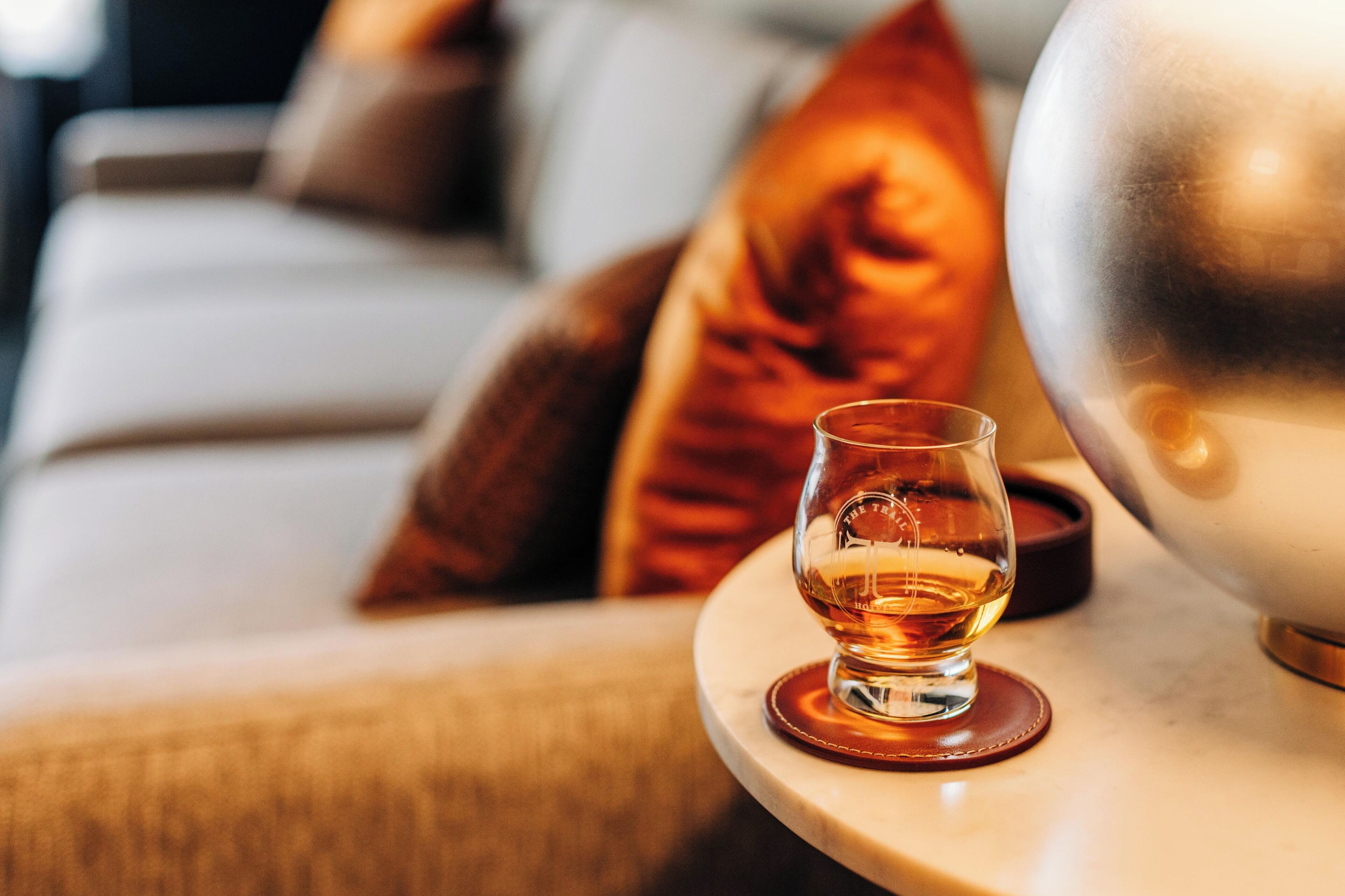 Ein Glas Whiskey auf einem Untersetzer neben einem metallischen Gegenstand, möglicherweise einer Lampe, auf einem Tisch in einem Zimmer mit einer Couch und Kissen.